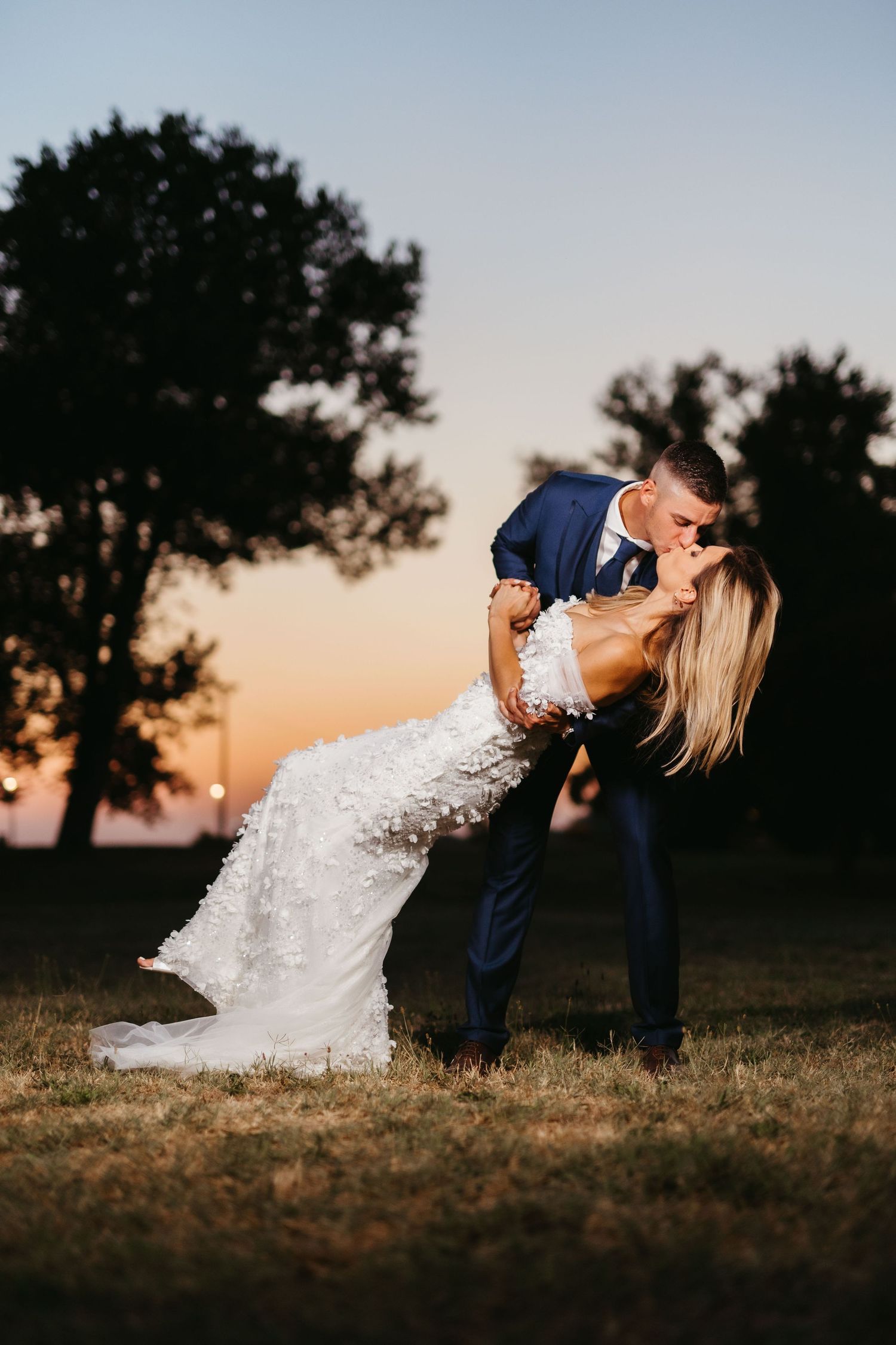 Dramatic wedding portrait features romantic dip pose against glowing sunset backdrop.