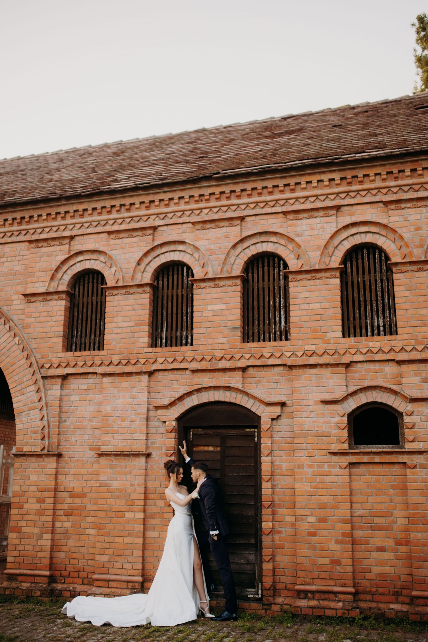 Elegant wedding couple walks along rustic brick wall with arched windows creating a romantic architectural backdrop.