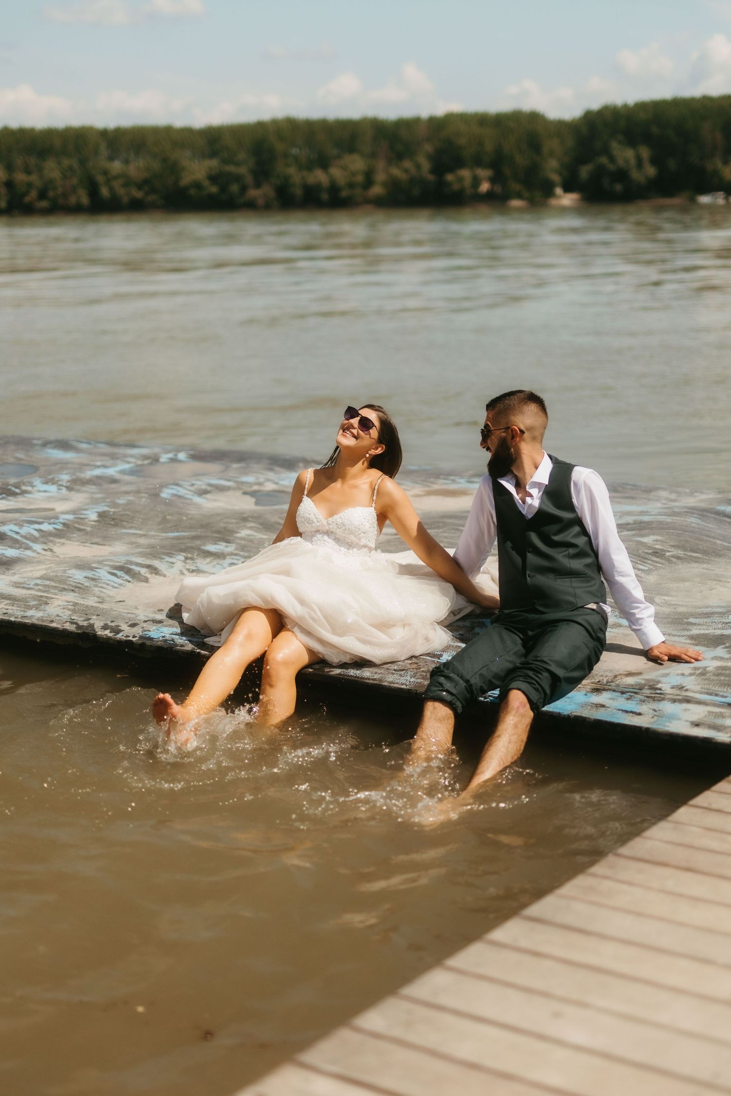 A couple in wedding attire sit on a dock with their feet in lake water on a sunny summer day.