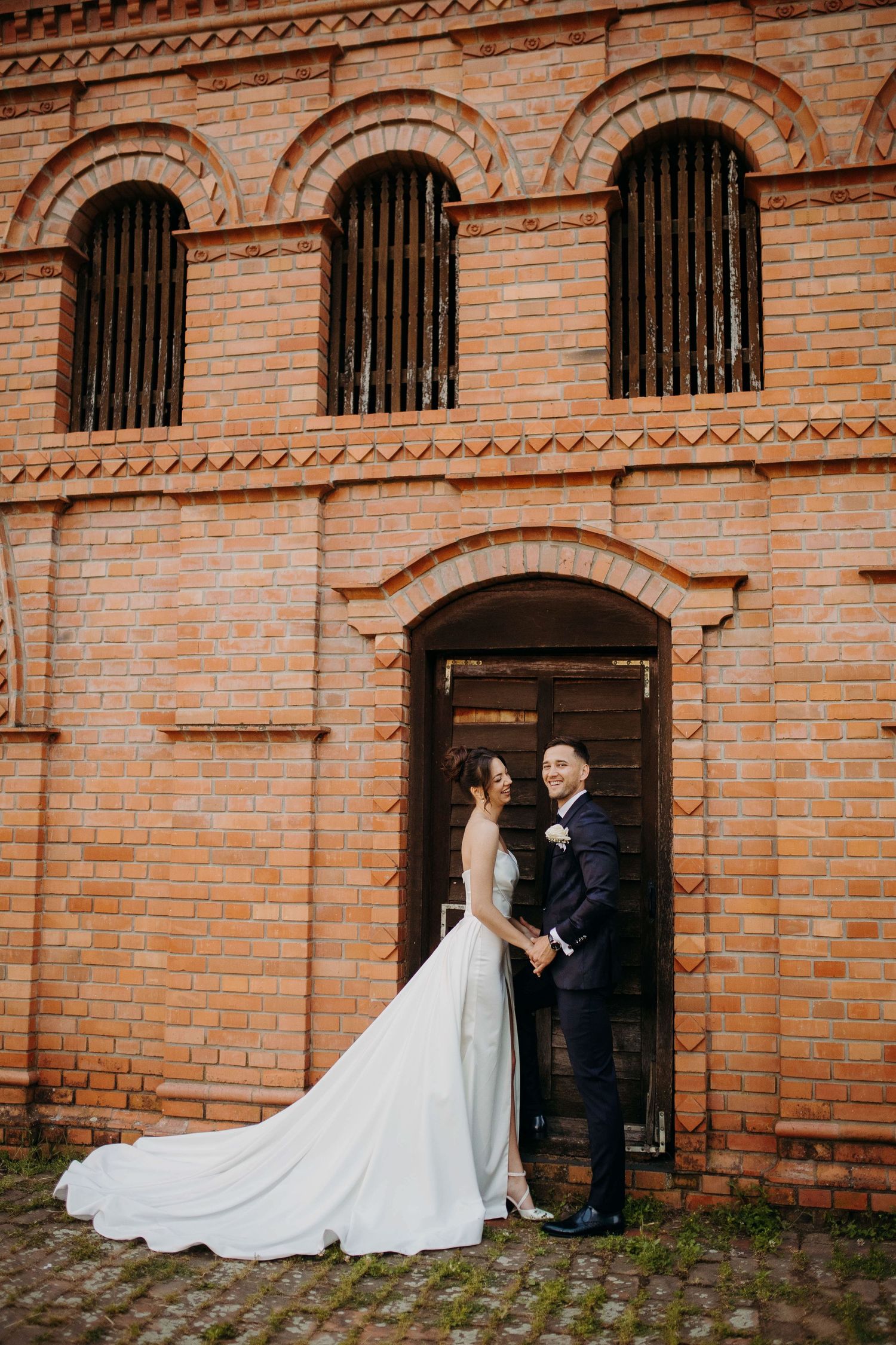 Elegant wedding couple stands in historic brick archway with ornate architectural details and iron-barred windows.