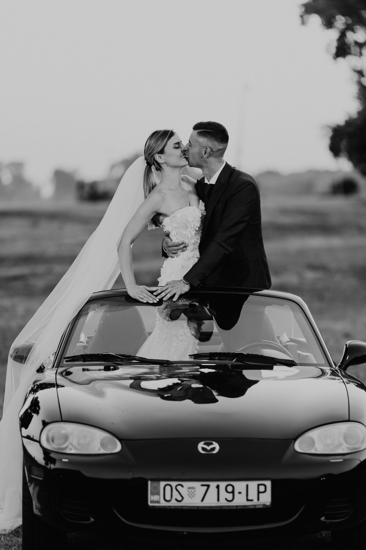 Black and white wedding portrait of couple embracing on hood of Mazda Miata.