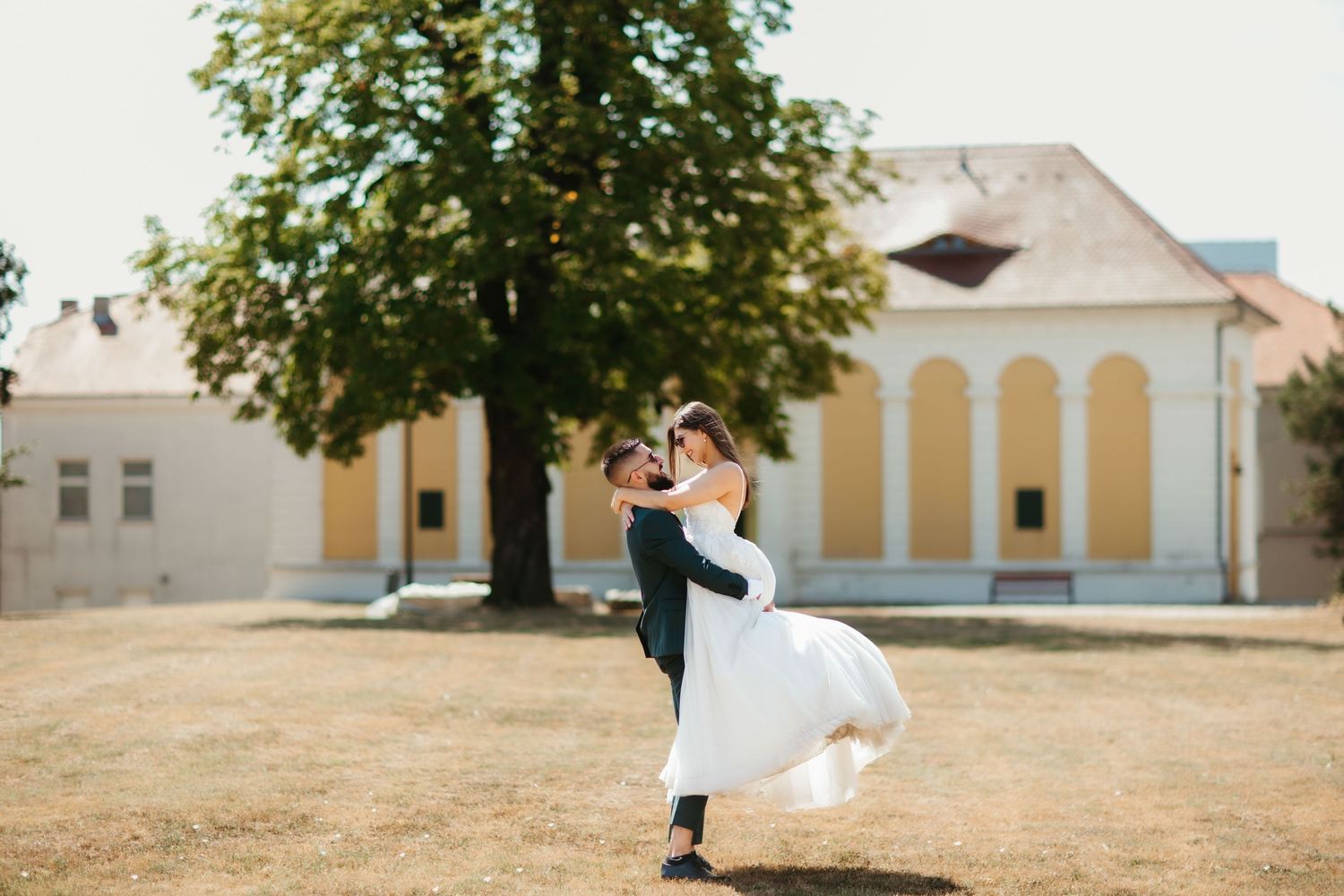 A romantic wedding couple embraces in front of historic white buildings on a sunny day, creating an elegant panoramic view.