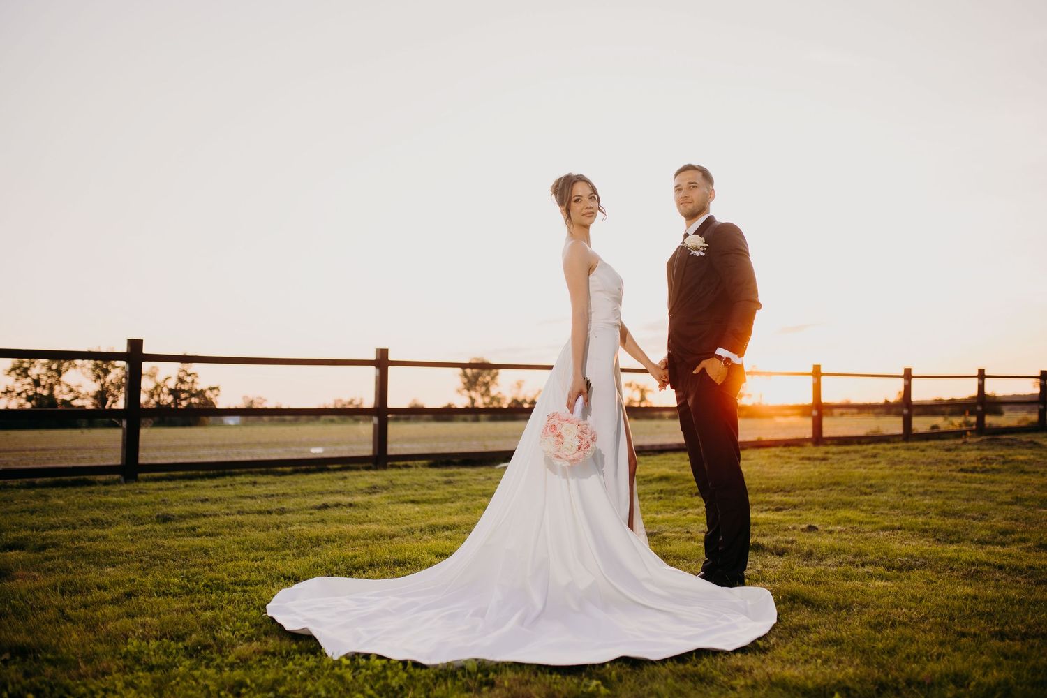 A couple poses for a sunset wedding photo on a grassy hilltop with a wooden fence in the background.