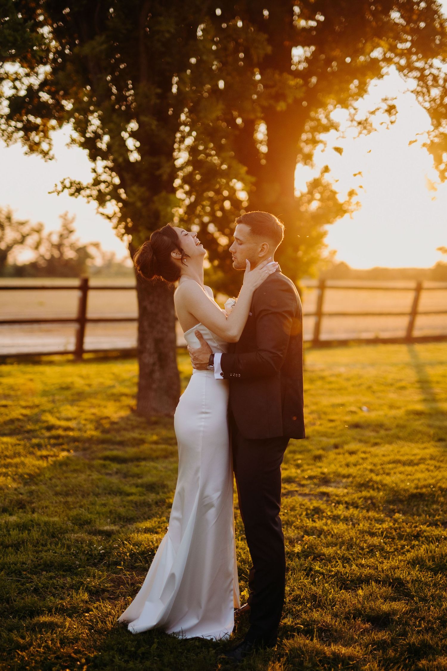 A romantic wedding couple embraces at sunset in a field with golden light streaming through trees and wooden fence.