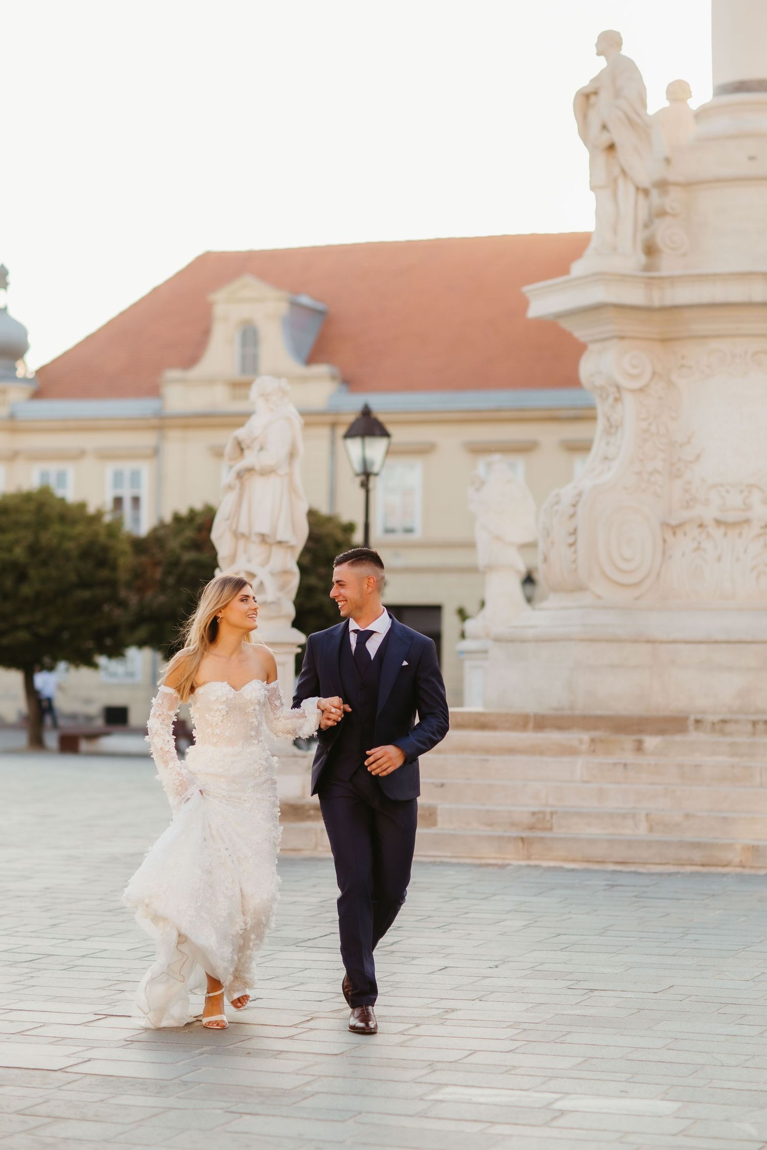 Couple walking together in formal attire near historic European architecture and statues.