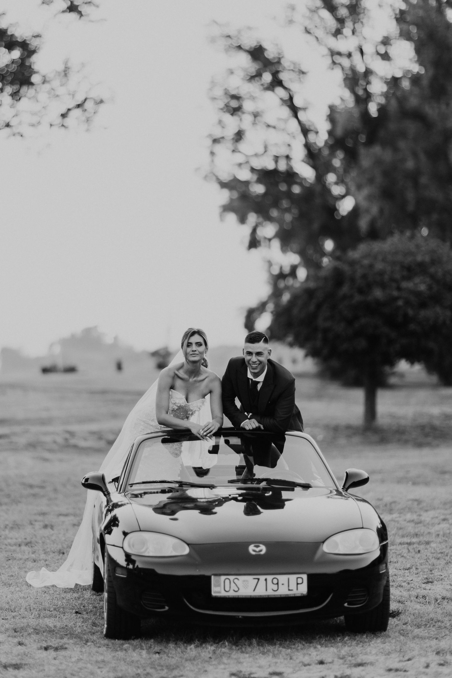 Black and white photo of newlyweds sitting on hood of classic Mazda Miata convertible.