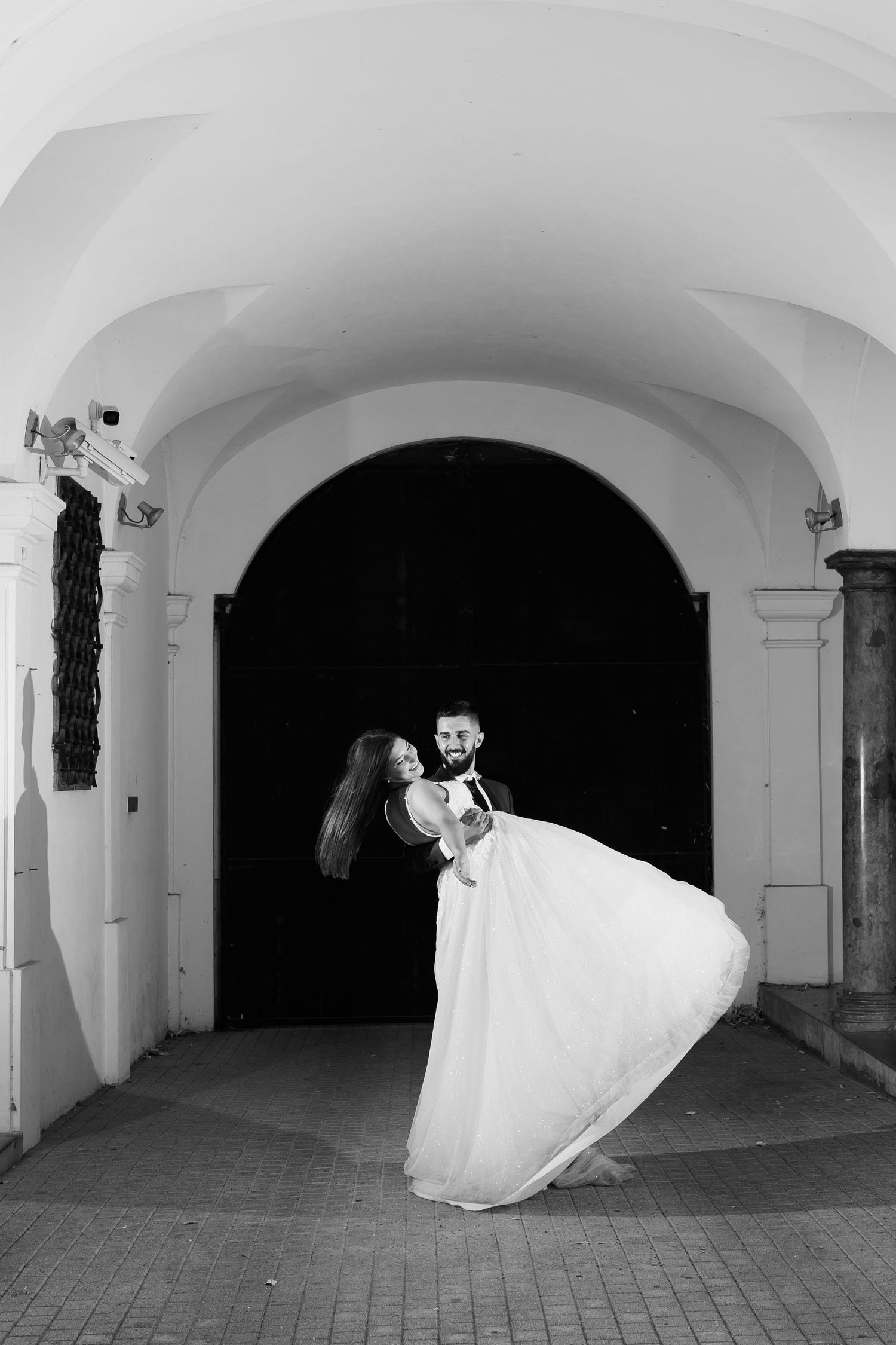 A romantic black and white wedding photo showing a couple in an elegant archway, with the bride's dress flowing dramatically.