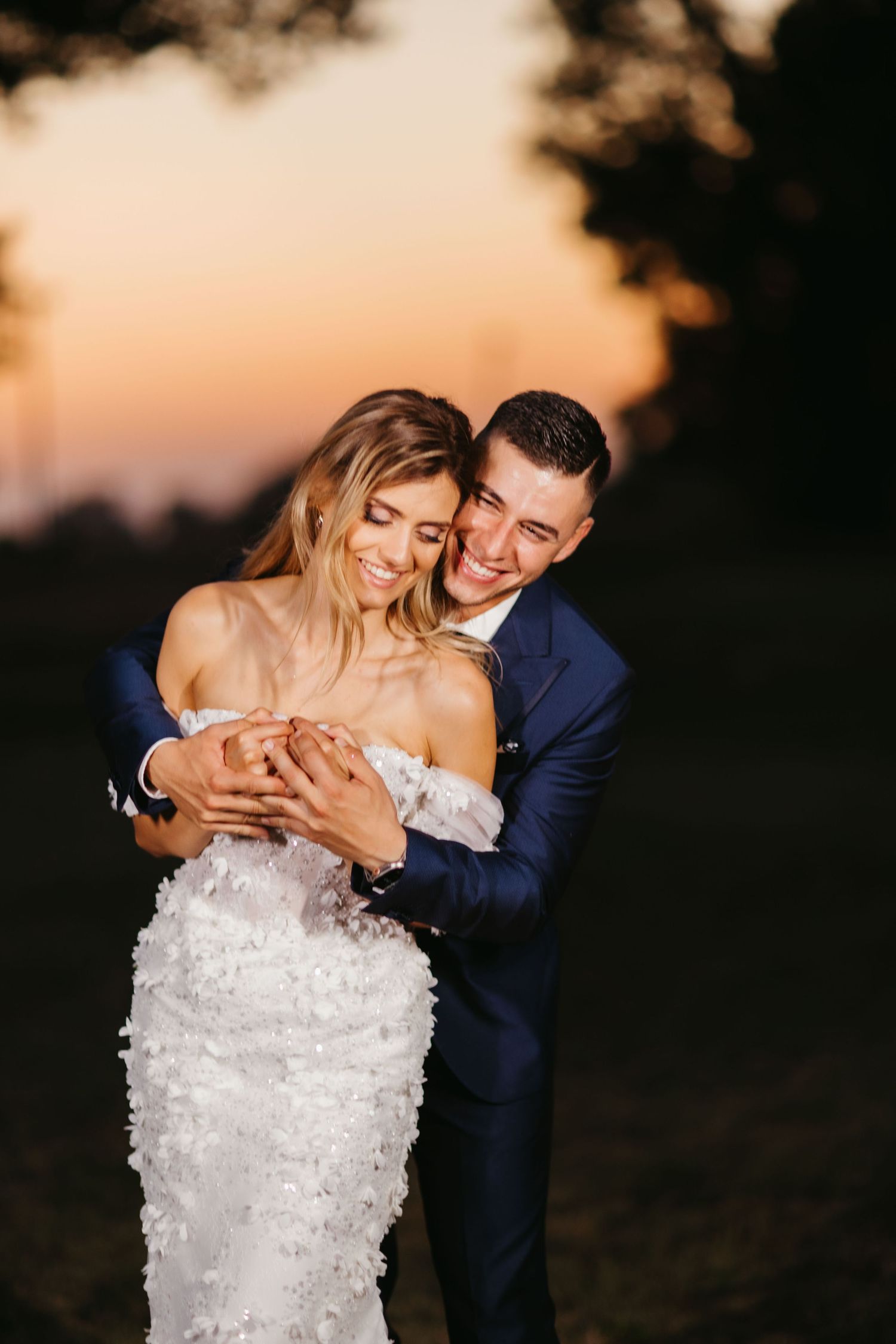 Romantic outdoor wedding photo of couple embracing at sunset with bride in white lace dress and groom in navy suit.
