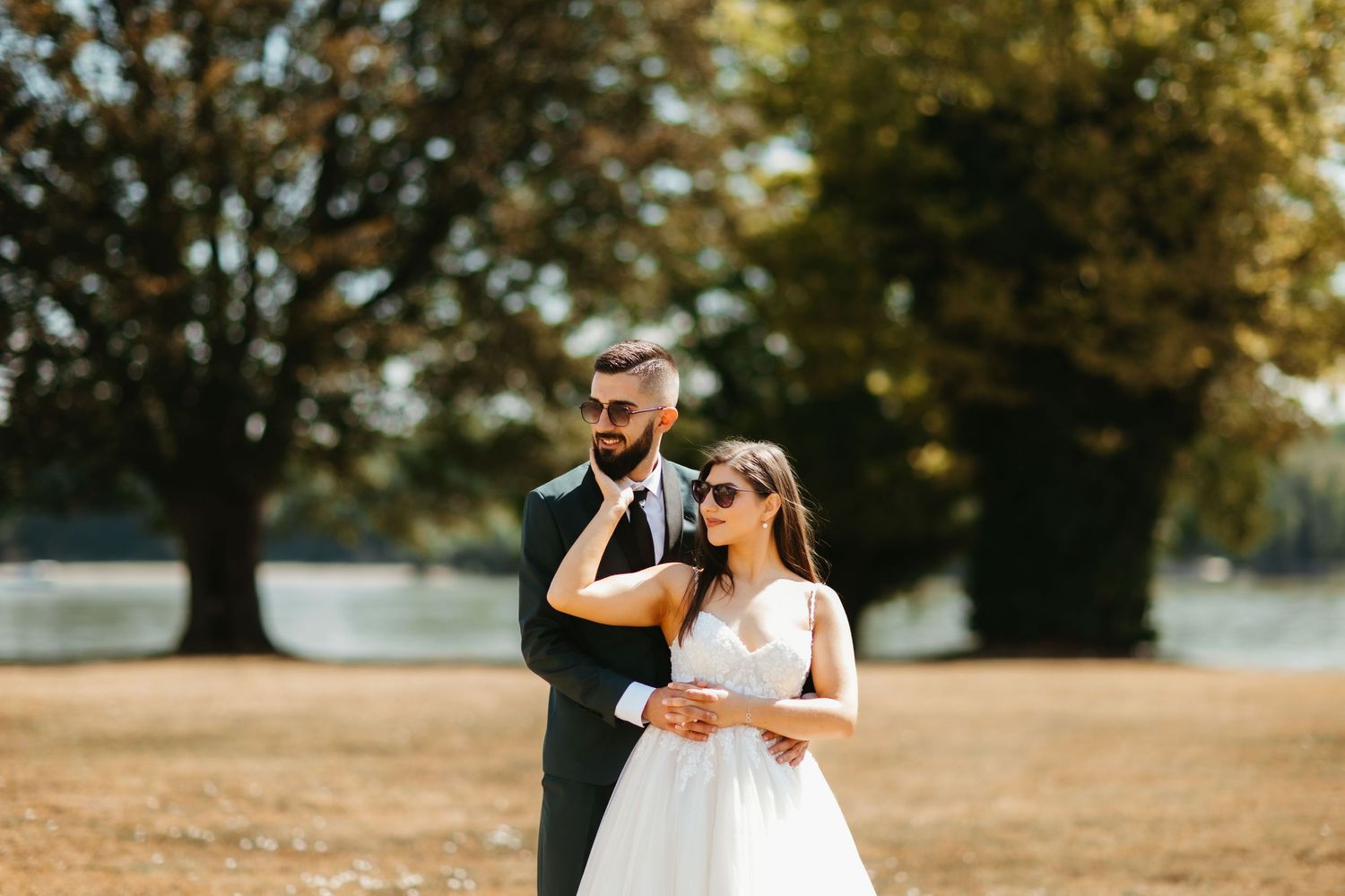 A happy couple laughs together on their wedding day in a scenic autumn park setting by a lake.
