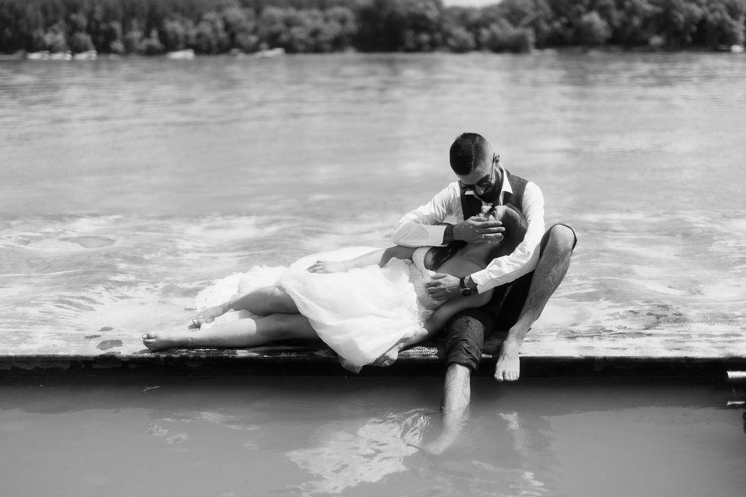 A romantic black and white photo shows a couple sitting together on a dock by the water.