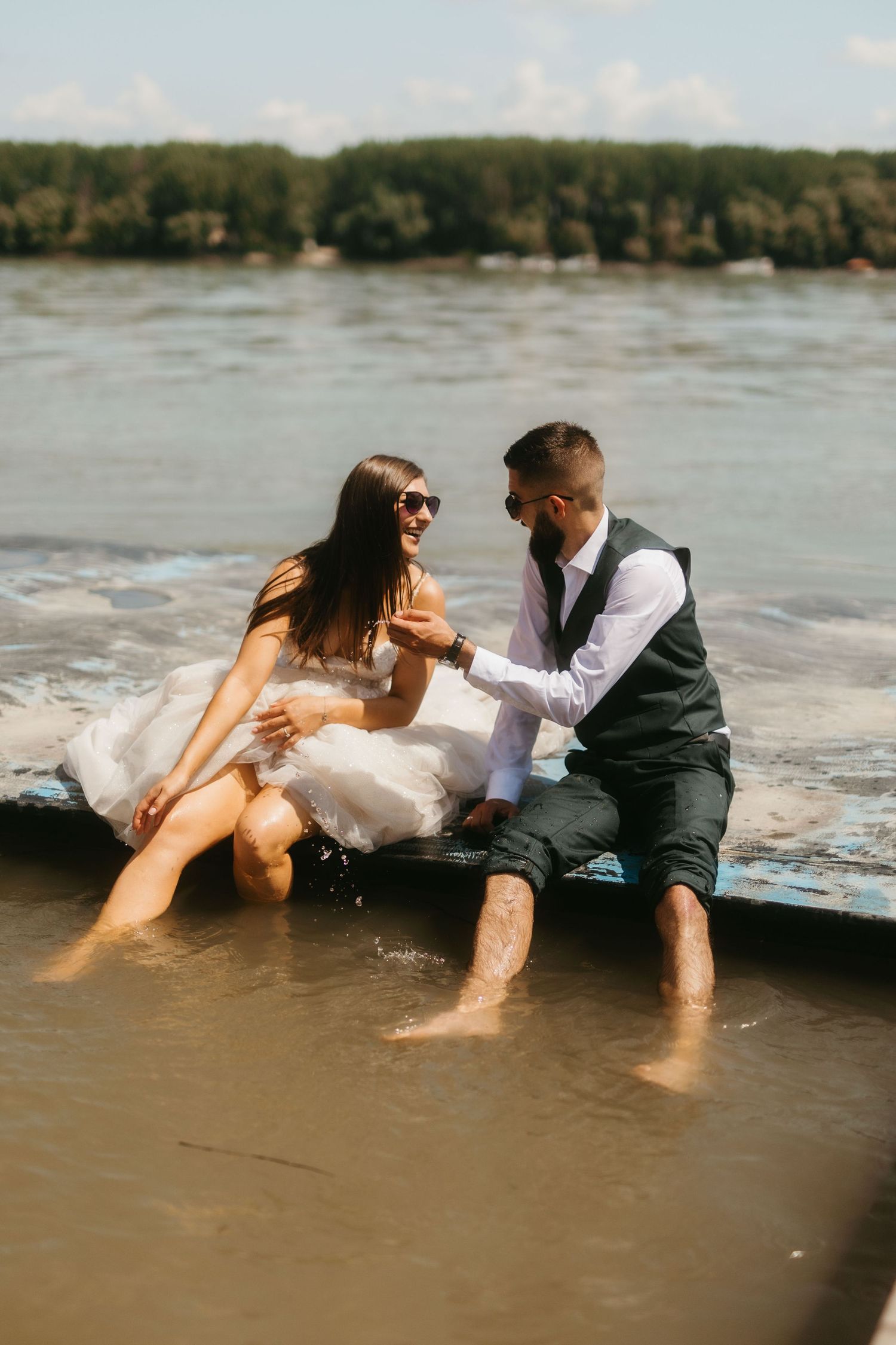 A couple in wedding attire sits at the edge of water, playfully dangling their feet in the river on a sunny day.