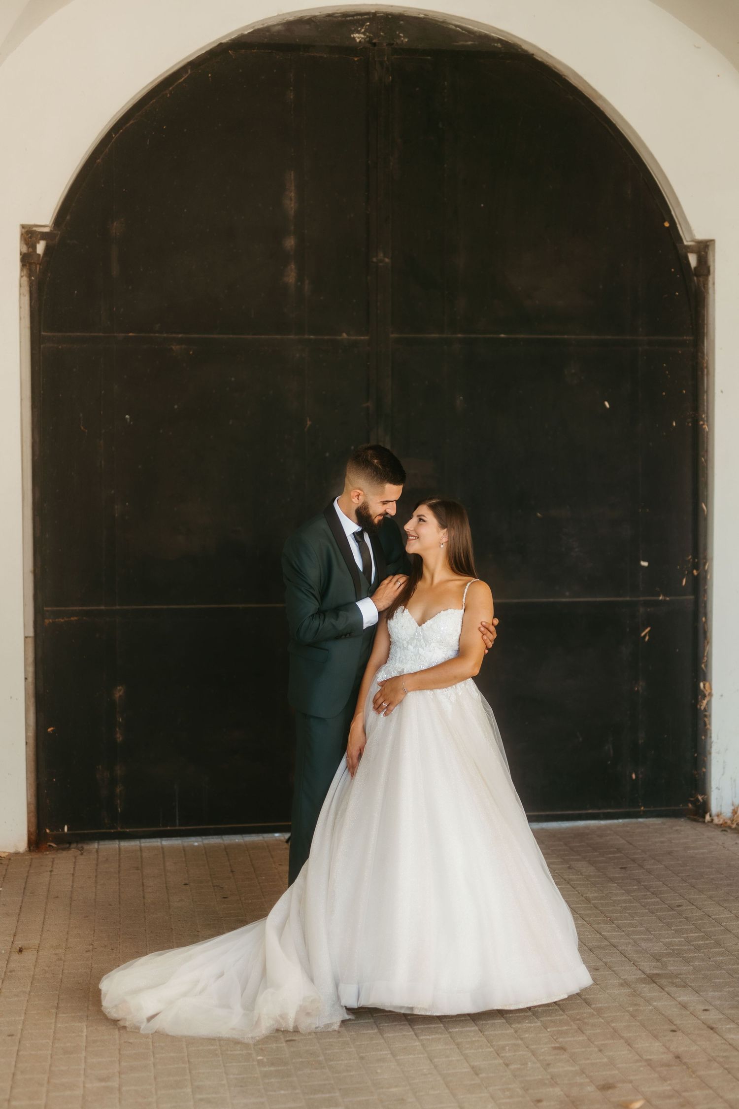 A couple embraces in wedding attire in front of a large arched dark door with warm vintage lighting.