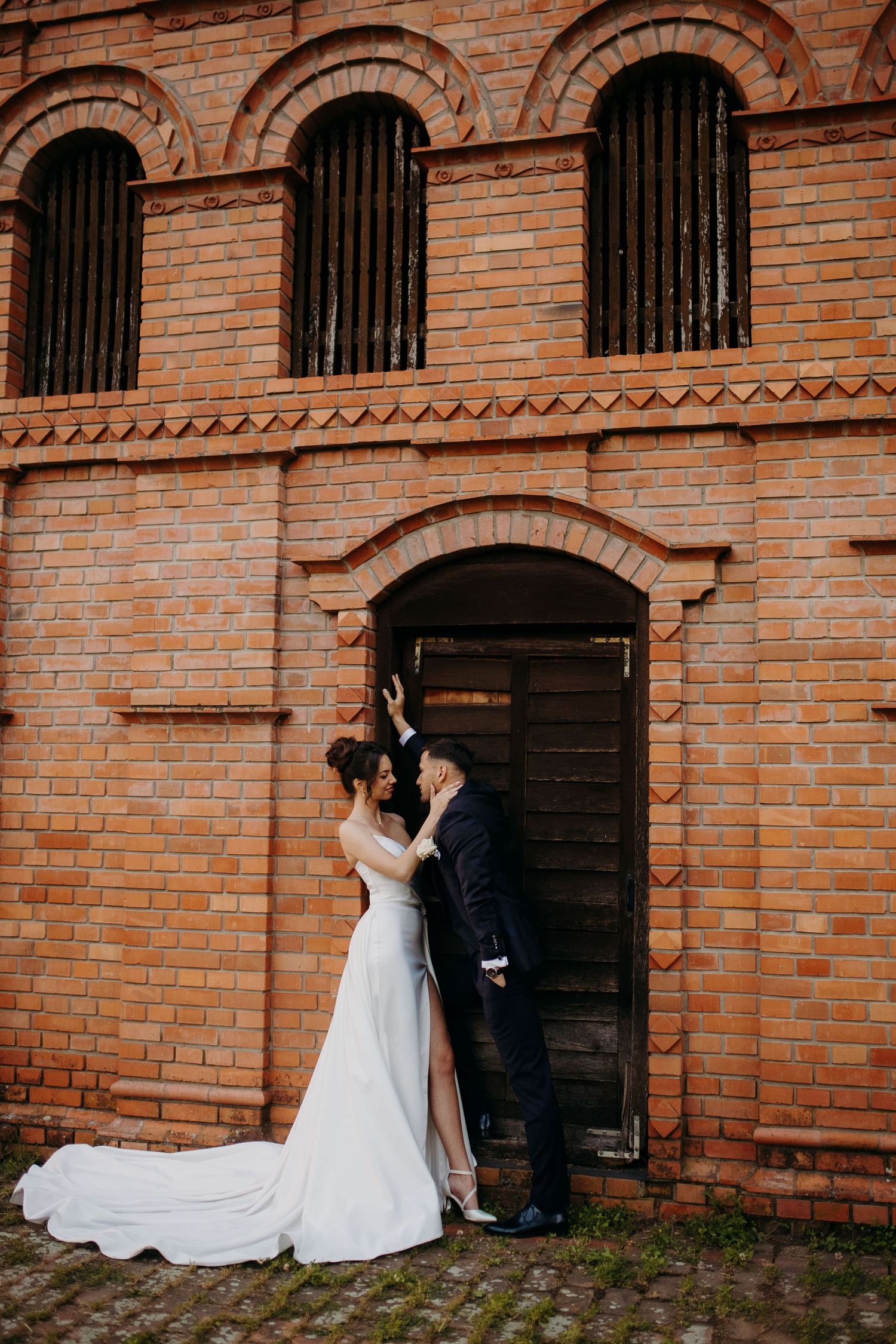 Couple in wedding attire embraces in front of rustic brick wall with arched windows and iron bars creating romantic ambiance.