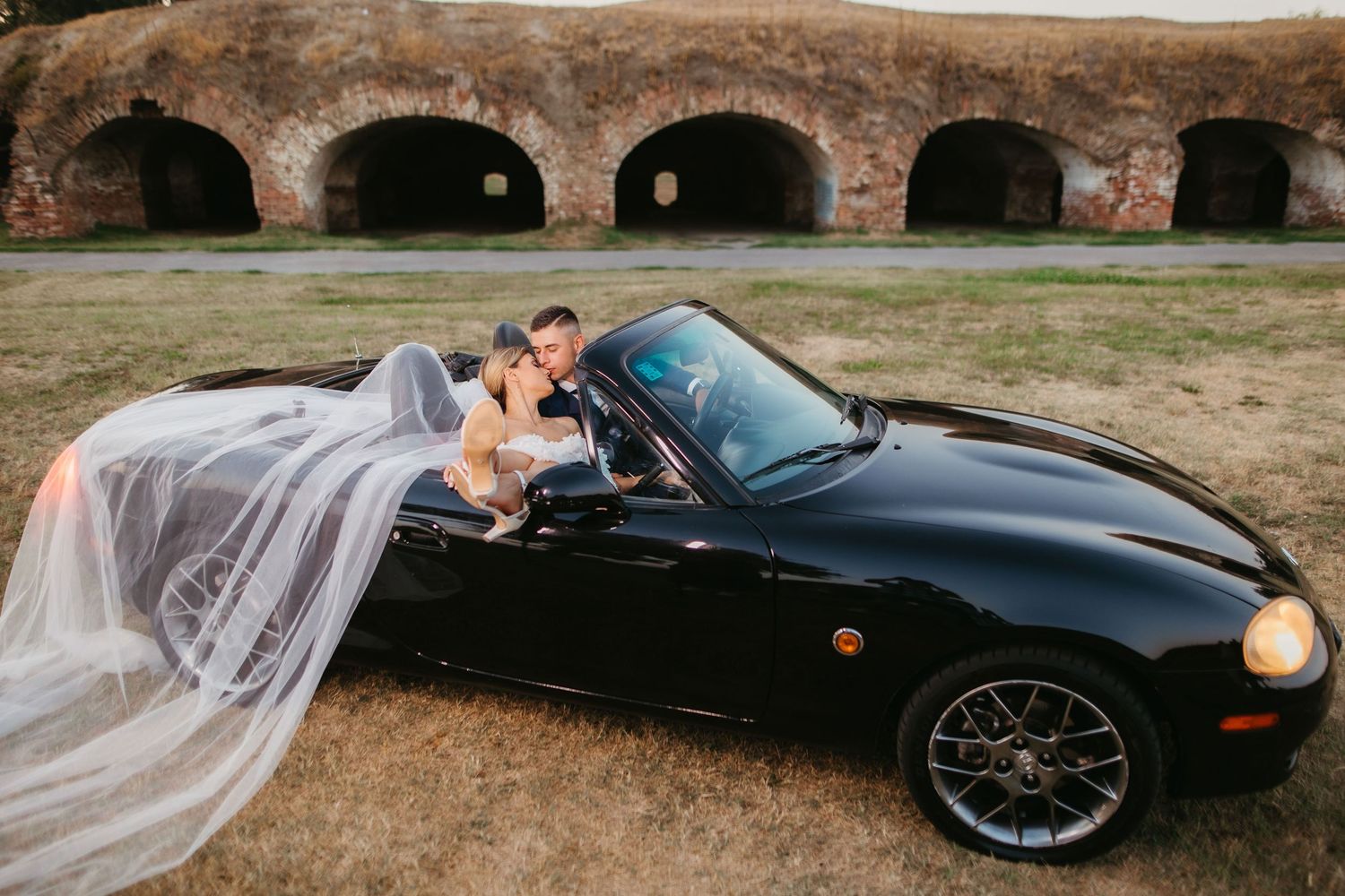 Romantic wedding shot of couple in black sports car with dramatic stone architecture.