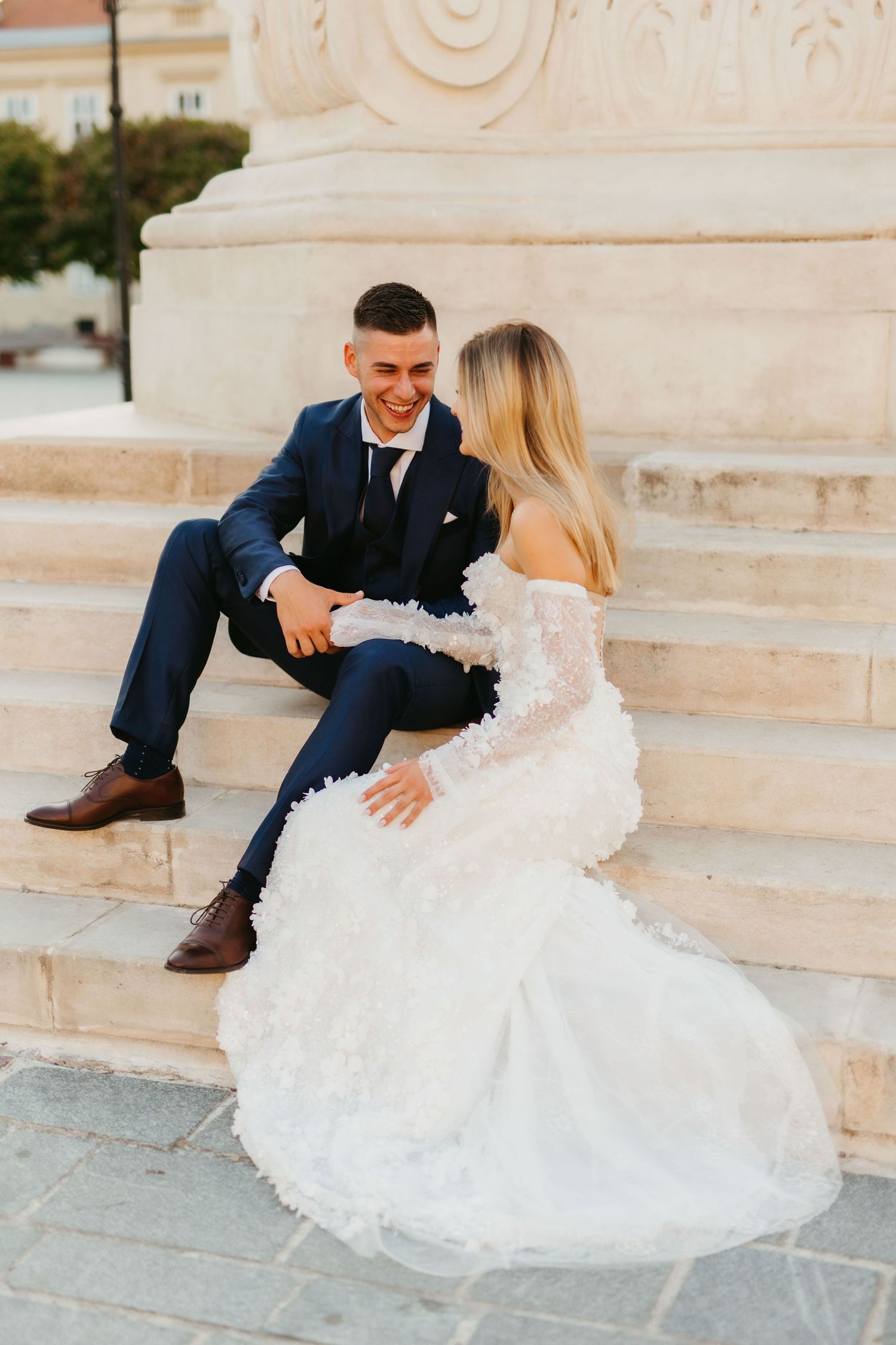 A couple in wedding attire sits casually on white marble steps.