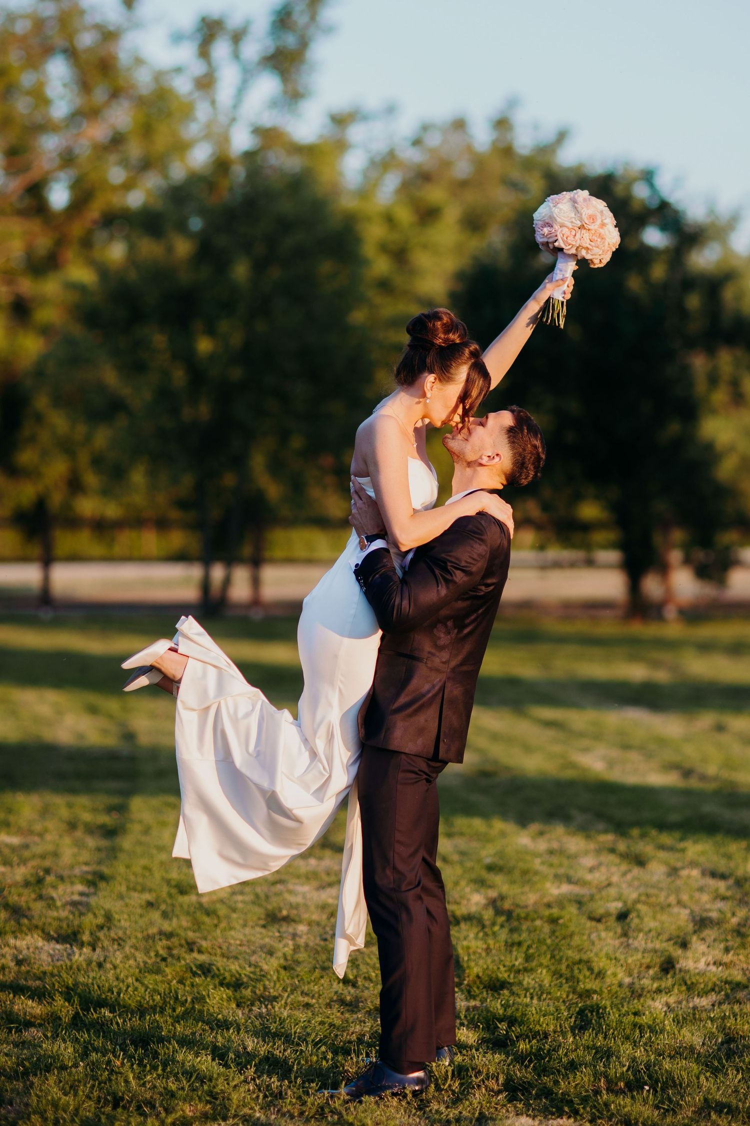 Newlyweds share romantic sunset moment in park as bride holds bouquet above while being lifted by groom.