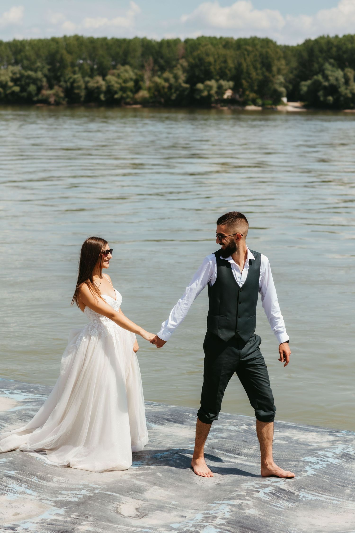 A couple stands barefoot in the waves along a tree-lined riverbank, with the person in a white dress holding hands with their partner.