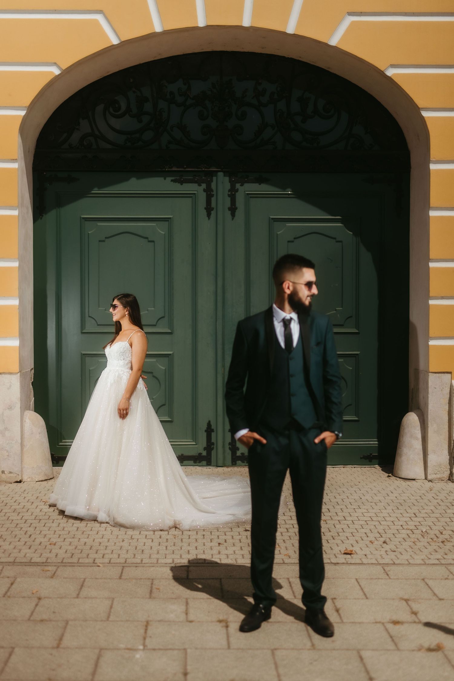 A couple in wedding attire pose apart against large green doors and yellow walls in a stylish architectural setting.