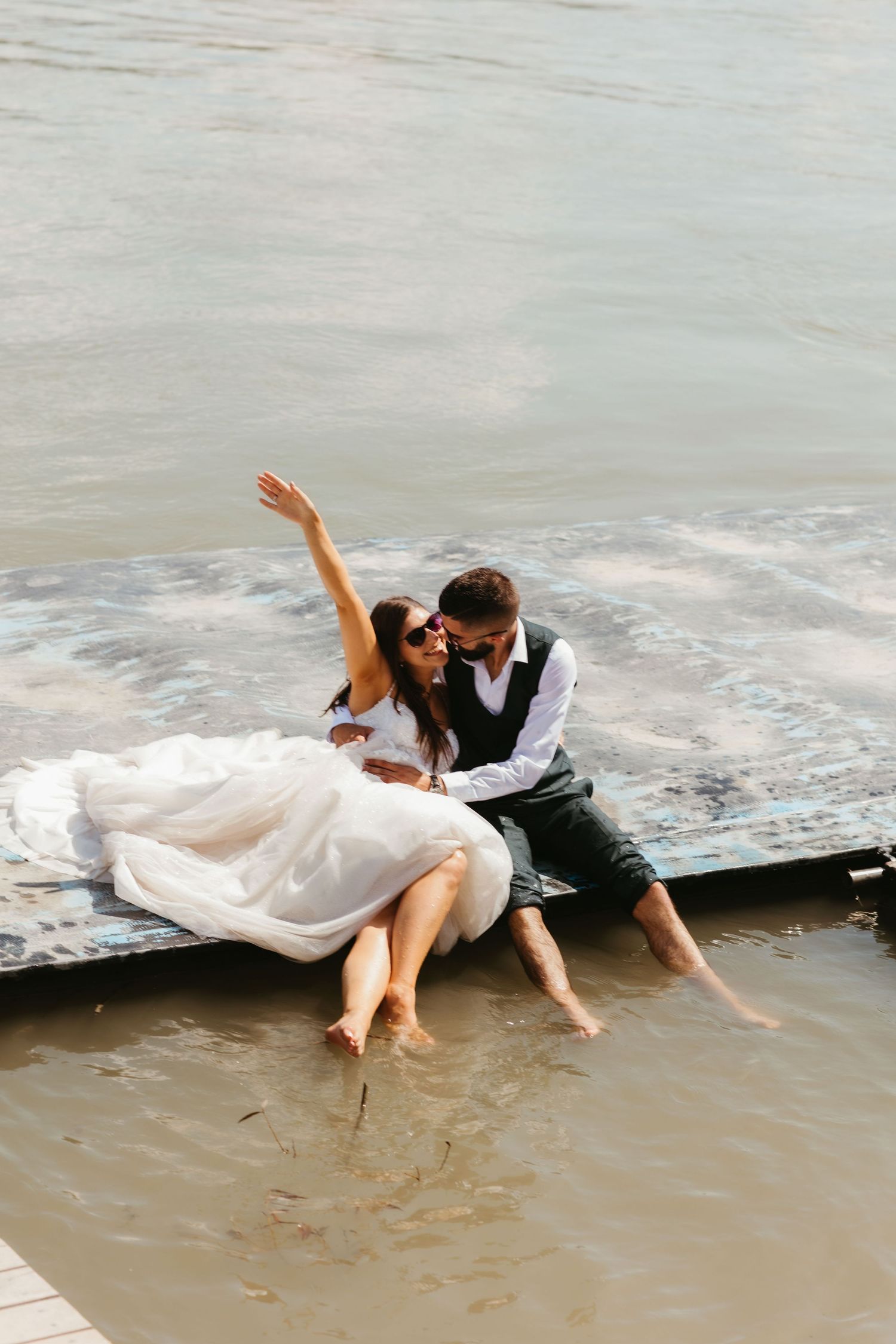 A couple in wedding attire playfully sits at the ocean's edge as waves crash around them on a sandy beach.