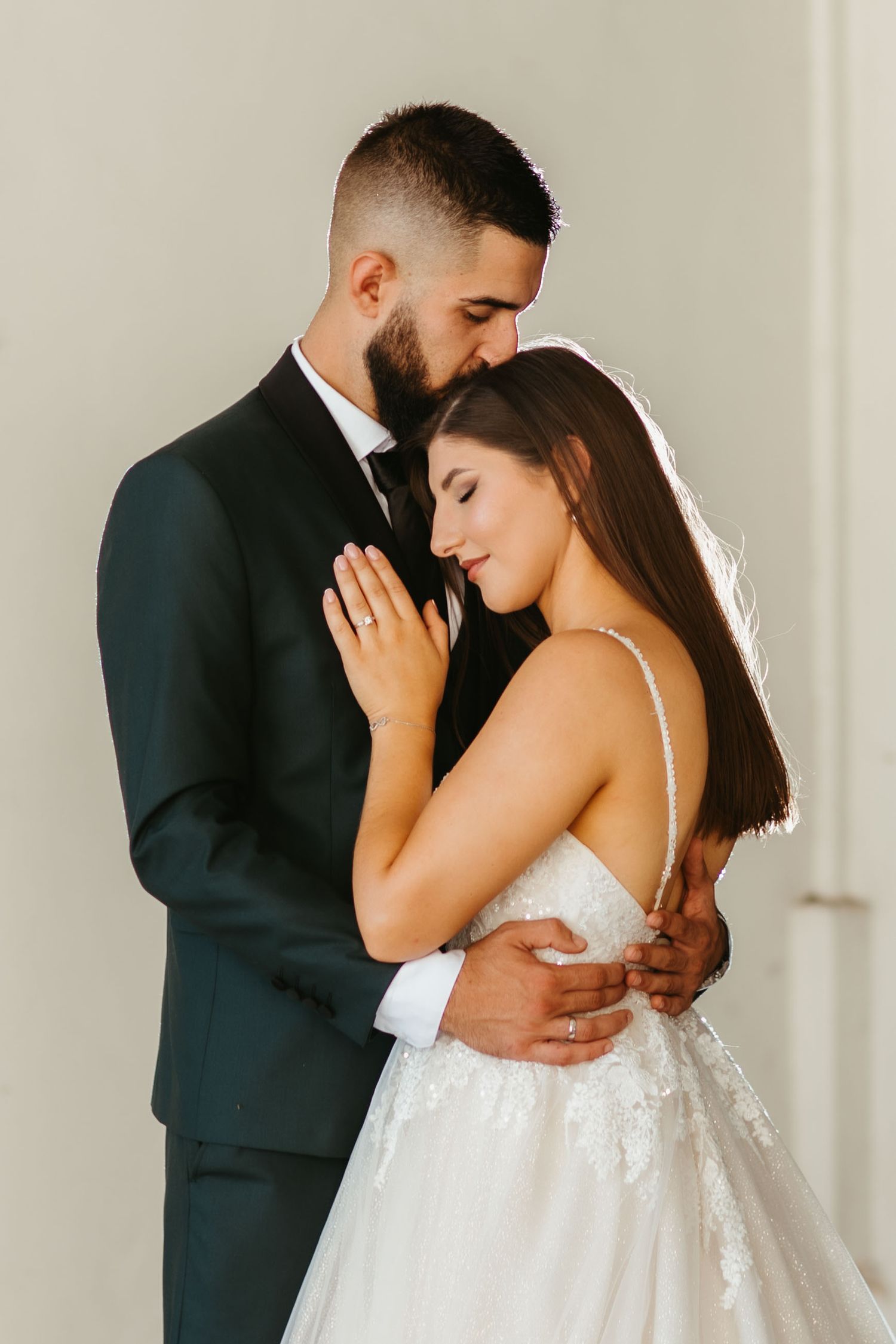 Romantic wedding portrait of couple embracing, bride in white lace gown and groom in dark suit against light background.