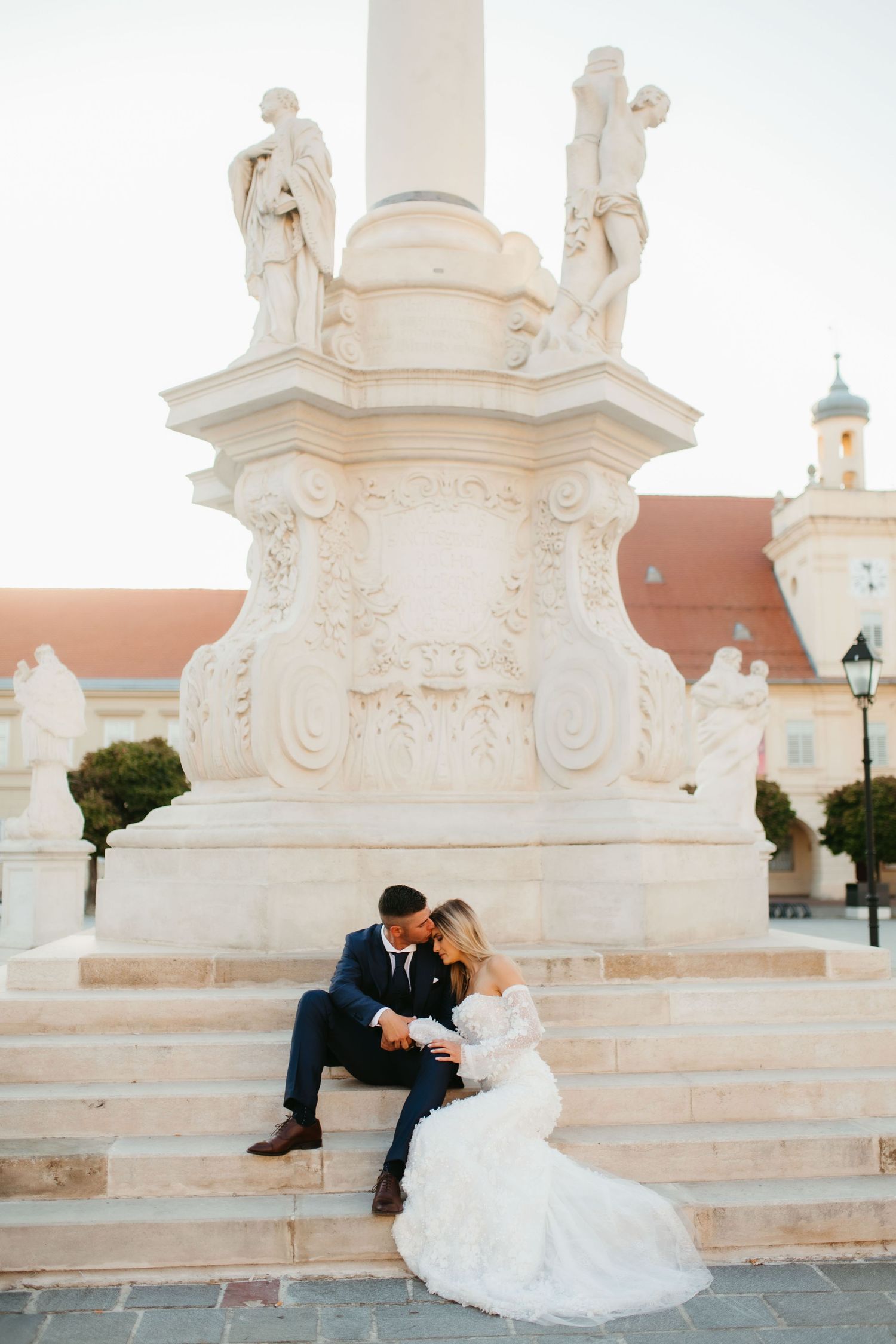 A romantic couple in formal attire share an intimate moment on ornate white stone steps at sunset.