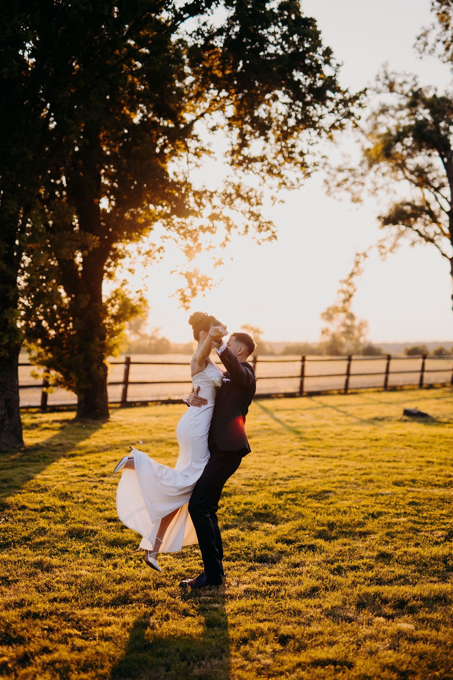 A couple dances and embraces in a golden sunset on grassy farmland with a wooden fence in the background.