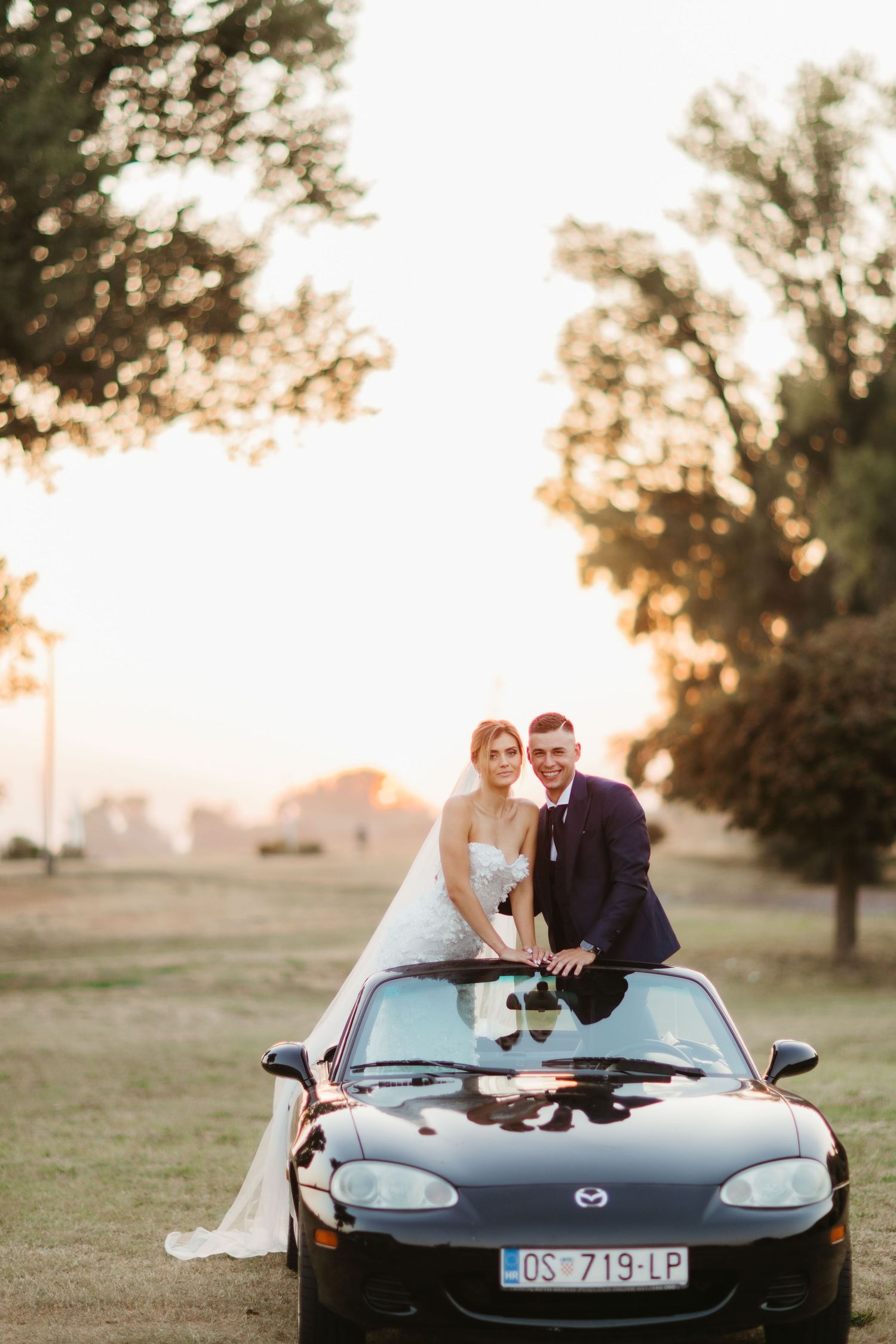 Wedding portrait with couple atop black Mazda Miata convertible in parkland setting at sunset.