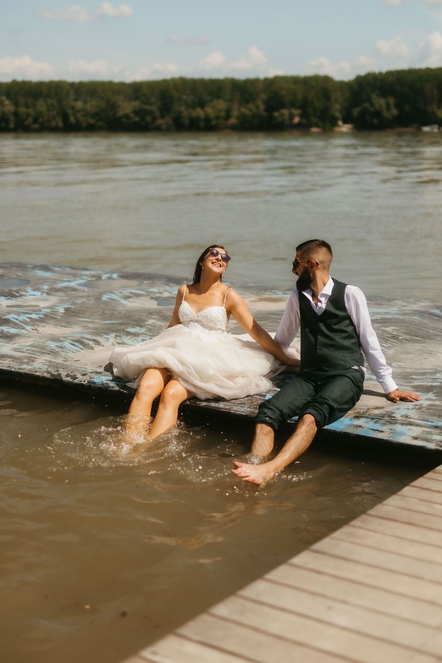 A couple sits at the edge of a dock, dangling their feet in the water while wearing wedding attire.