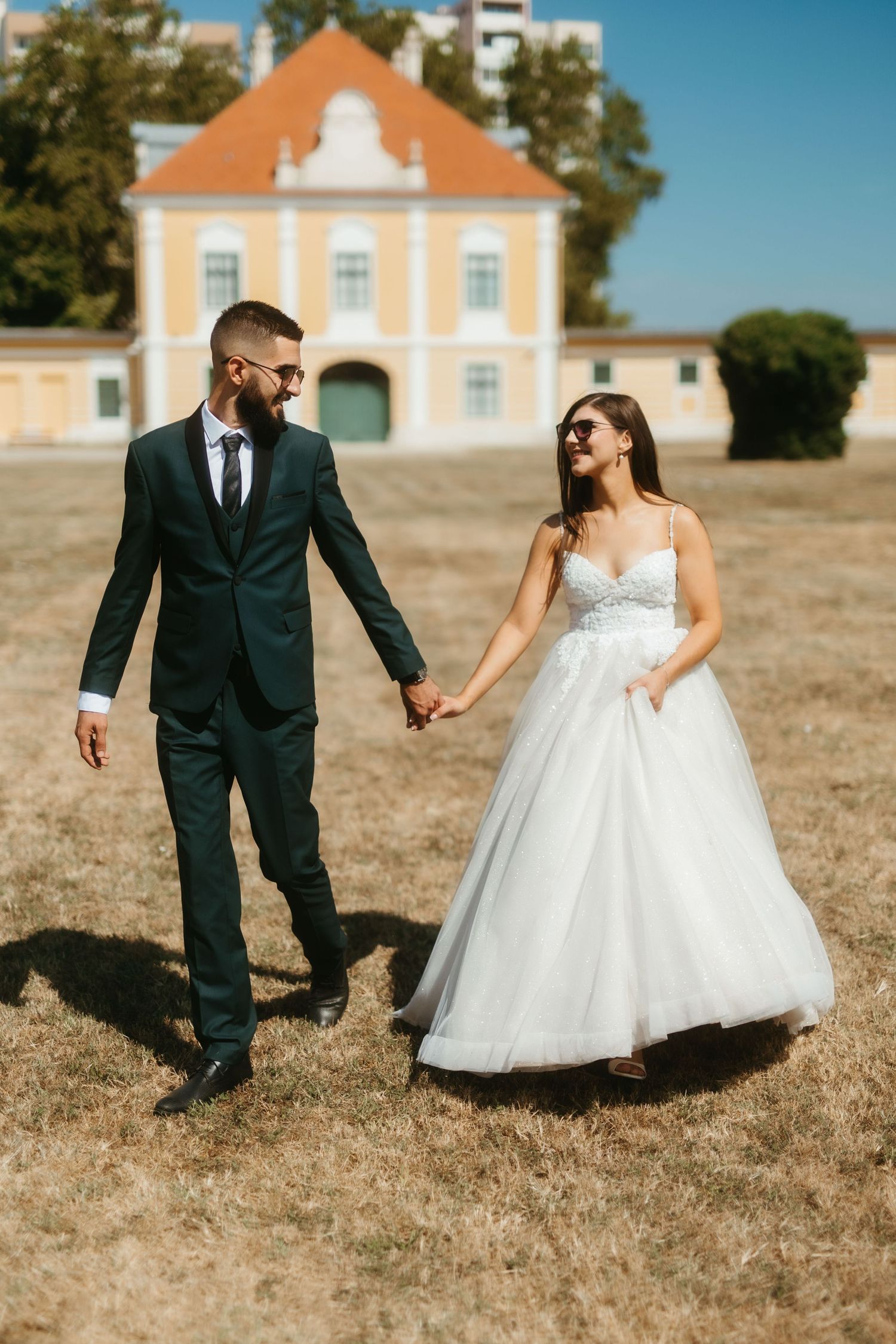 Couple in wedding attire walking hand in hand on dry grass field with orange-roofed historic building in background.