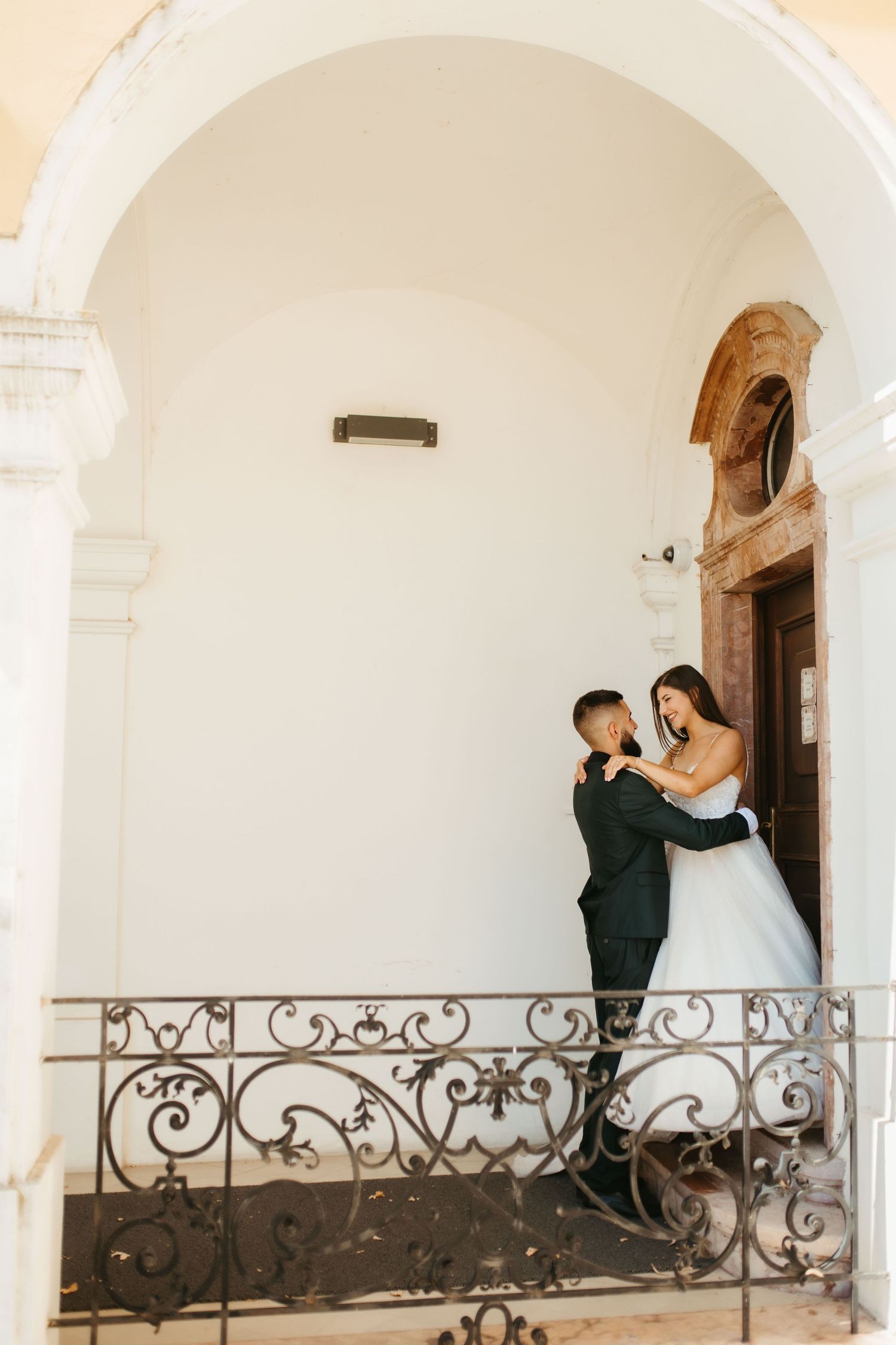 A bride and groom share a romantic moment under a white archway with ornate iron railings at their wedding.