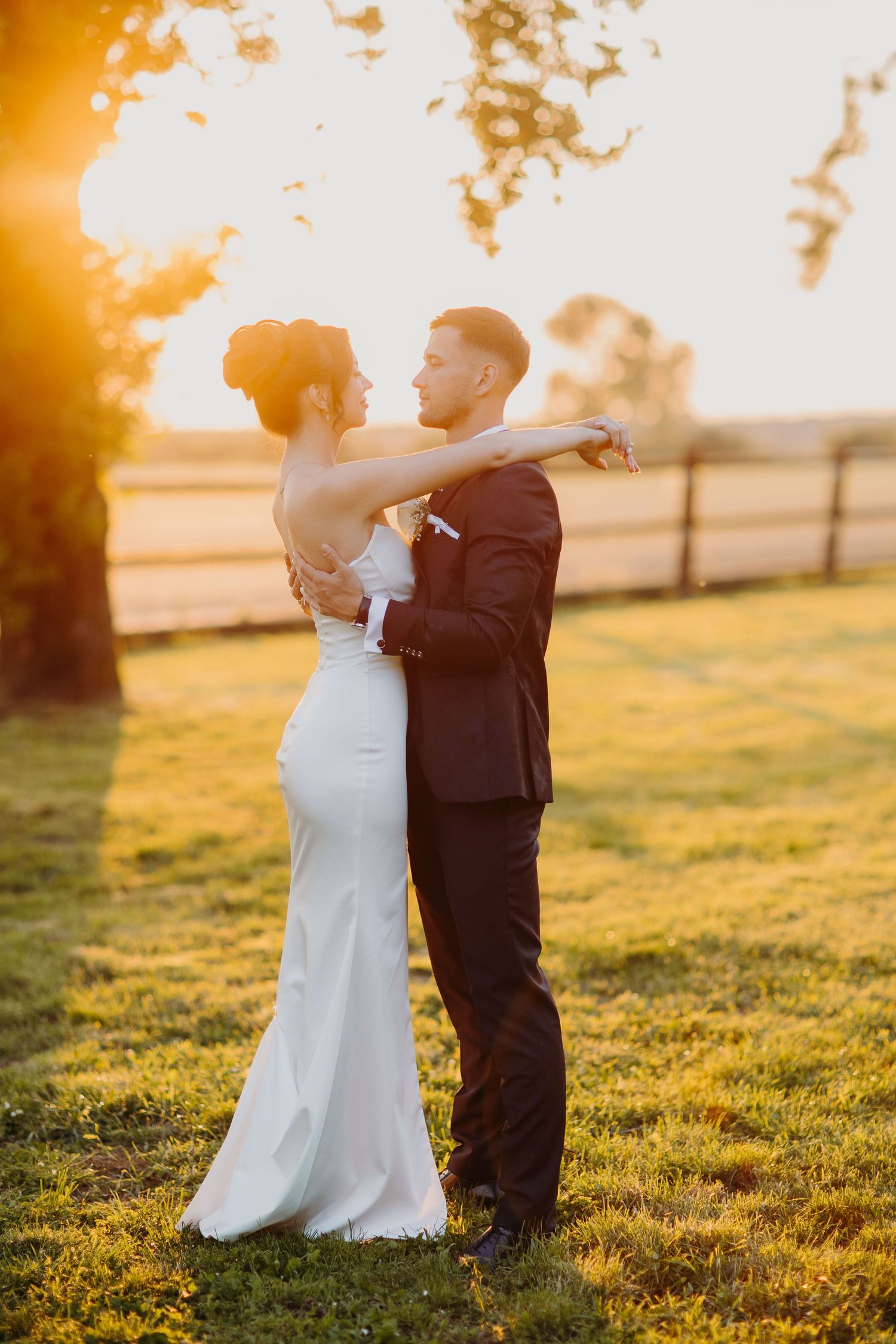 A wedding couple embraces during golden hour sunset on grassy field with soft warm lighting and romantic atmosphere.