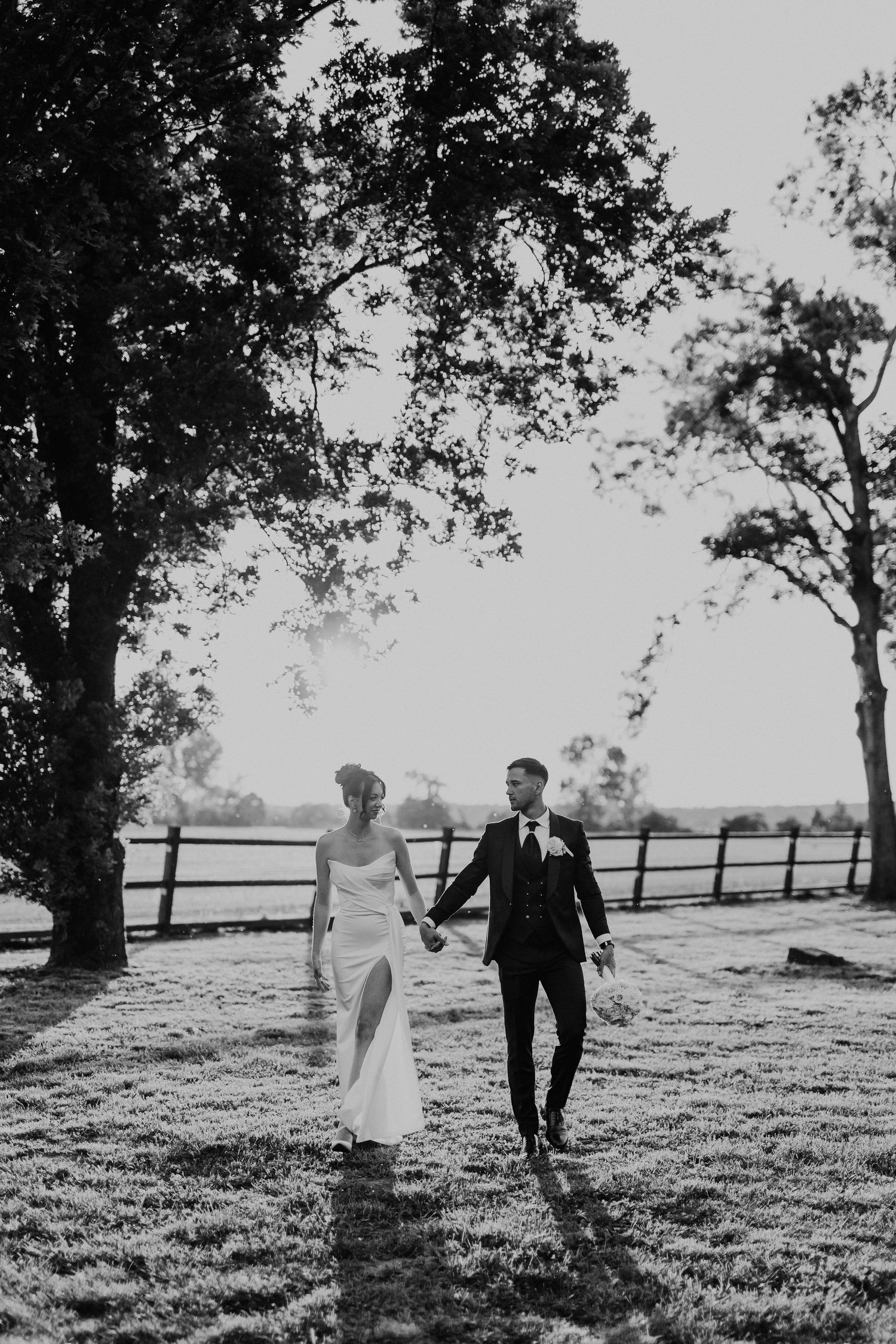 A black and white outdoor wedding photo of a couple walking hand in hand through a sunlit field with wooden fencing.