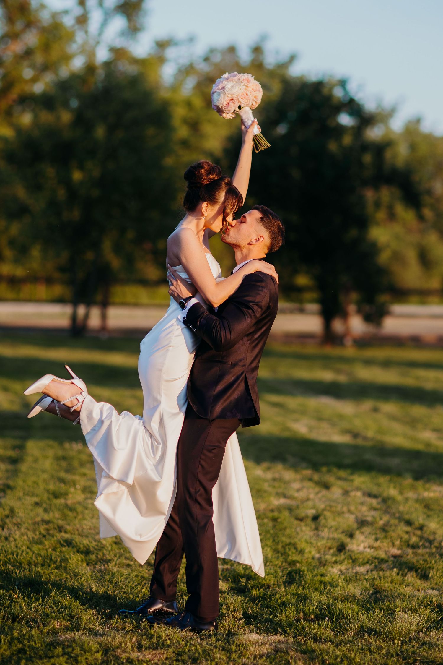 Bride and groom share a romantic lift during sunset photos, with the bride holding a pink bouquet against a golden sky.