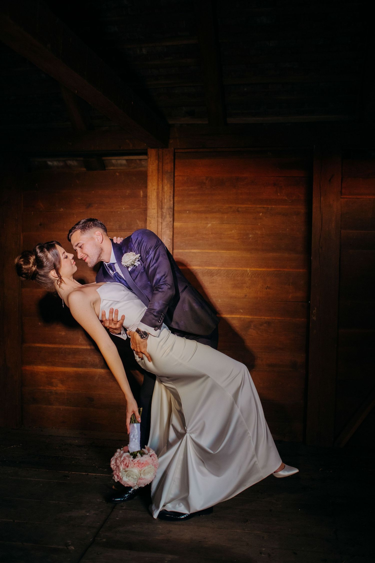 Romantic dancing couple in elegant attire share an intimate dip pose against wooden panel walls in moody lighting.