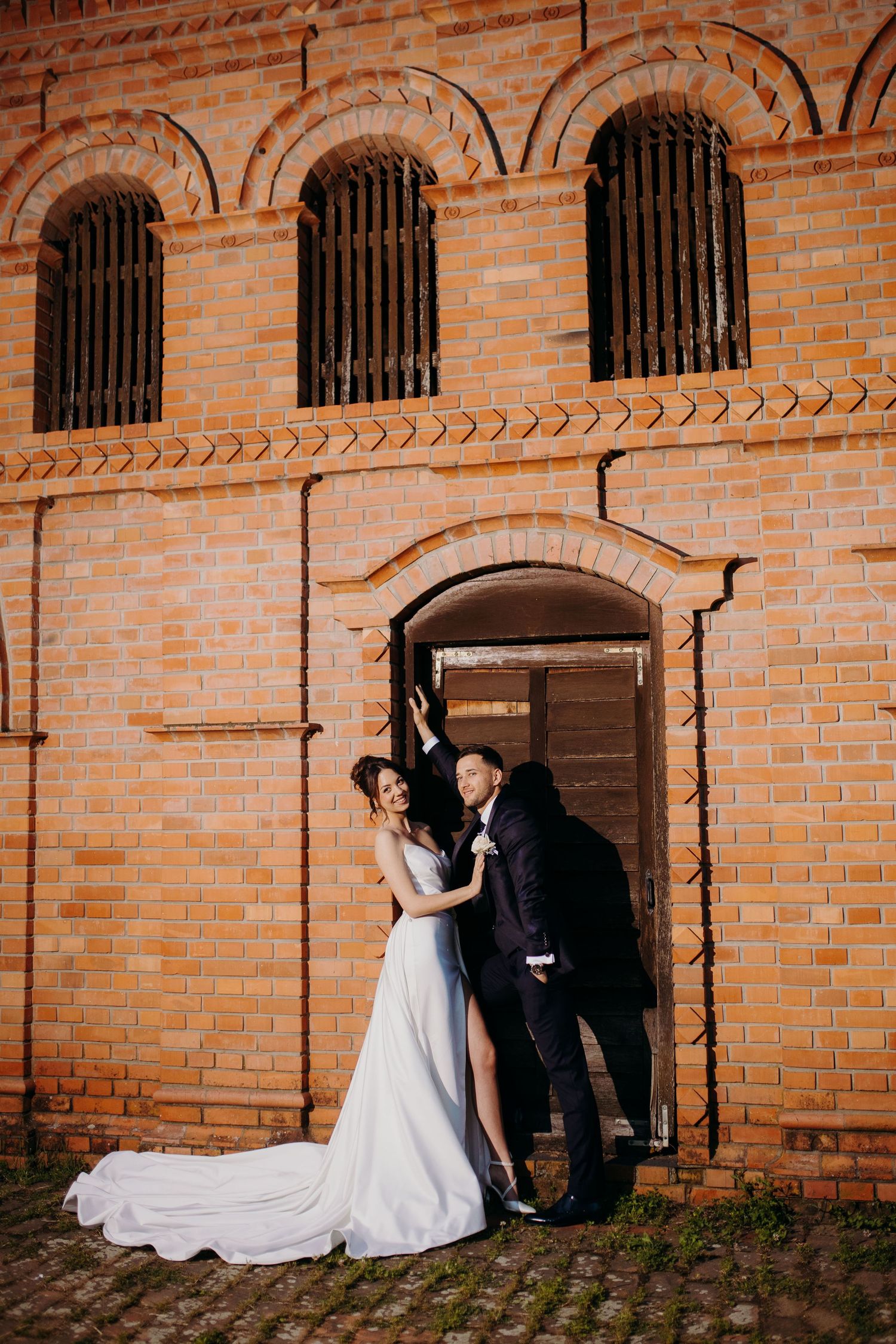 A romantic couple in wedding attire pose in an elegant brick archway of a historic building with arched windows.