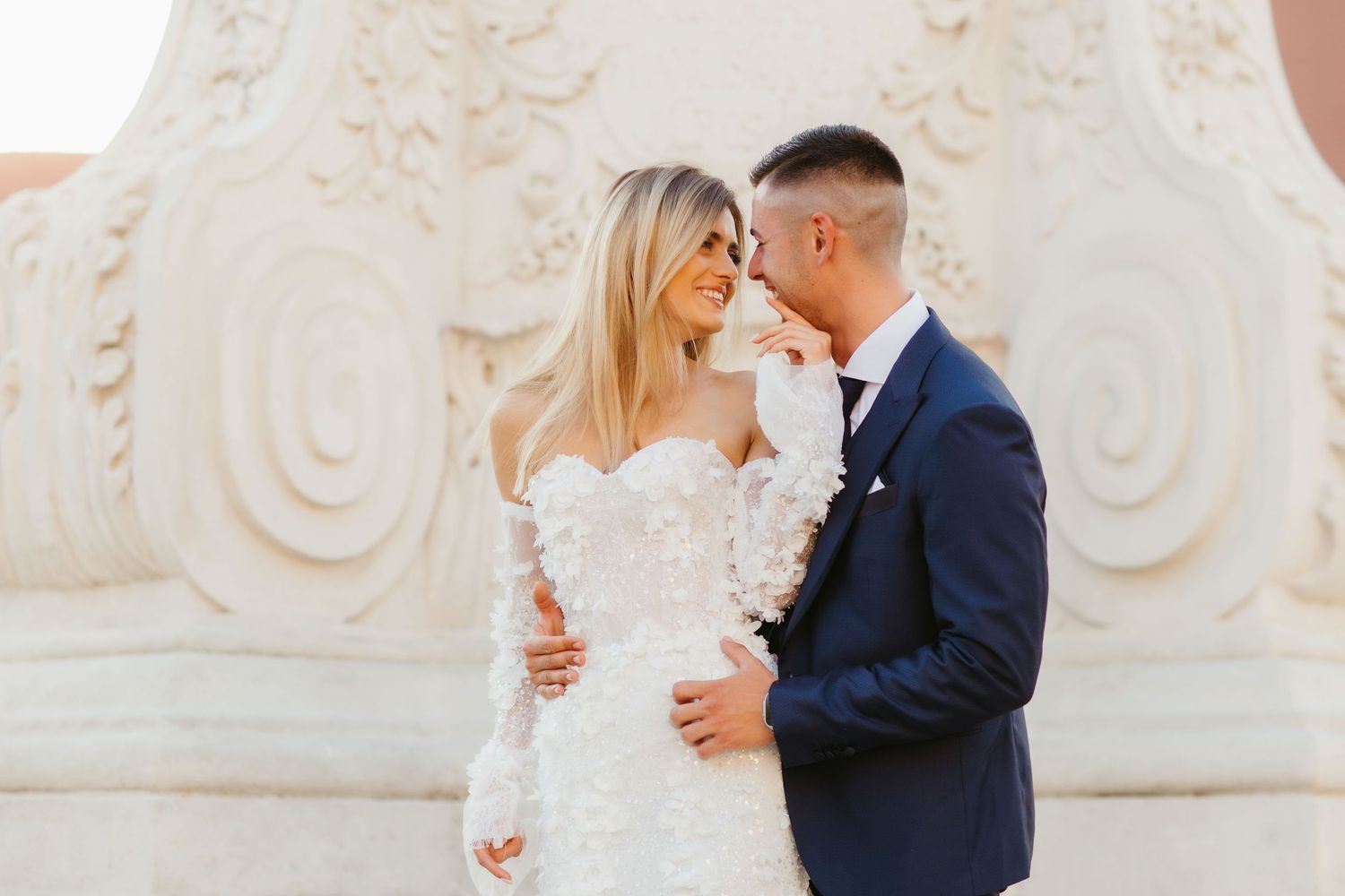Wedding couple in navy suit and white lace dress pose against ornate stone pillar.