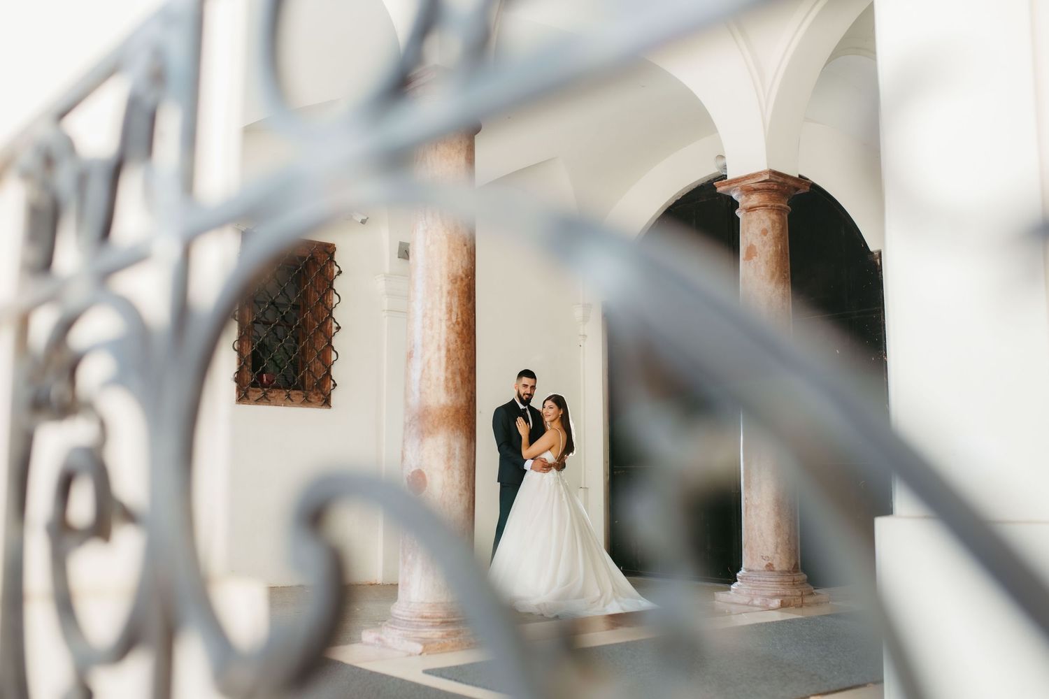 A romantic wedding couple embraces between elegant stone columns, viewed through ornate iron railings.