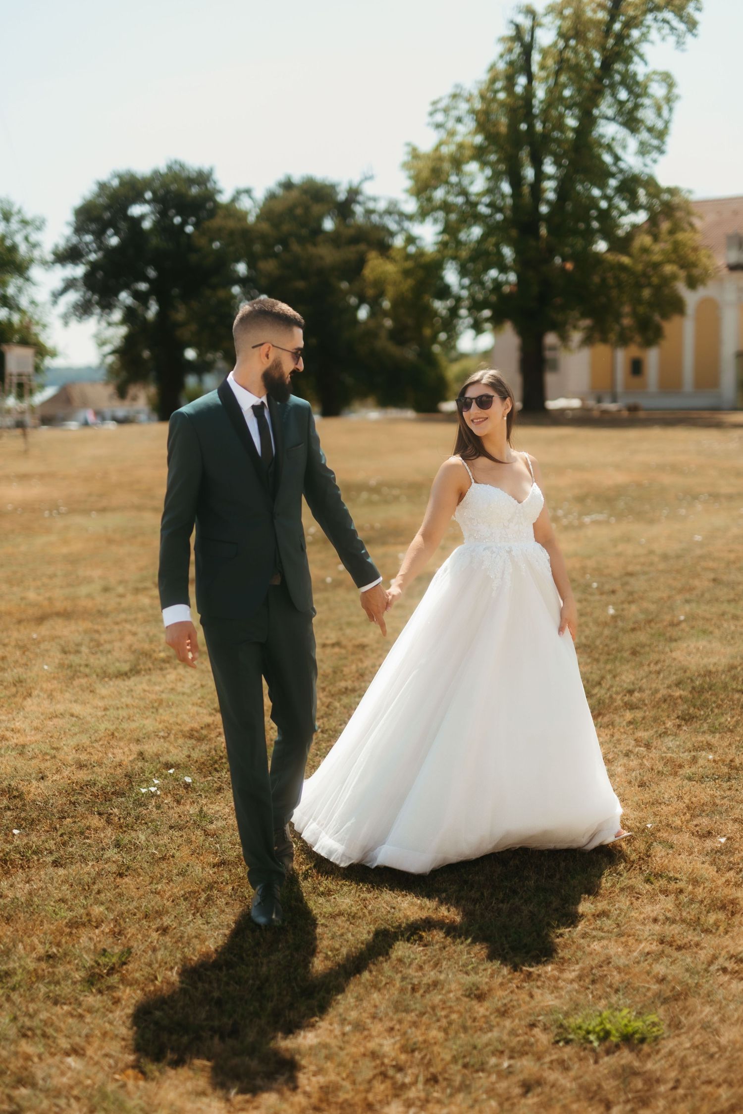 Bride and groom walk hand in hand across a sun-drenched golden field in elegant wedding attire.