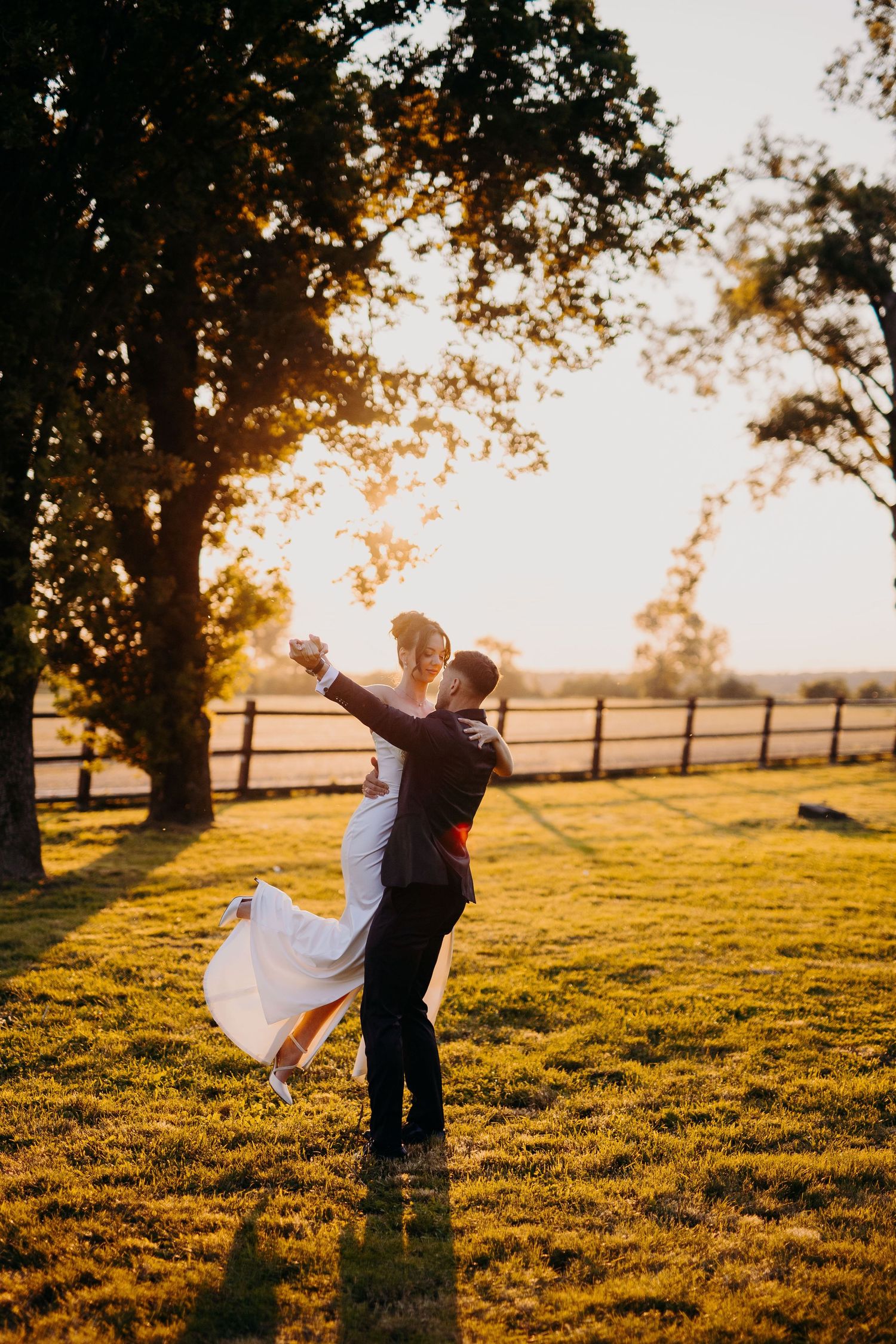A romantic couple embraces in a golden sunset on a grassy field with a wooden fence and trees in the background.
