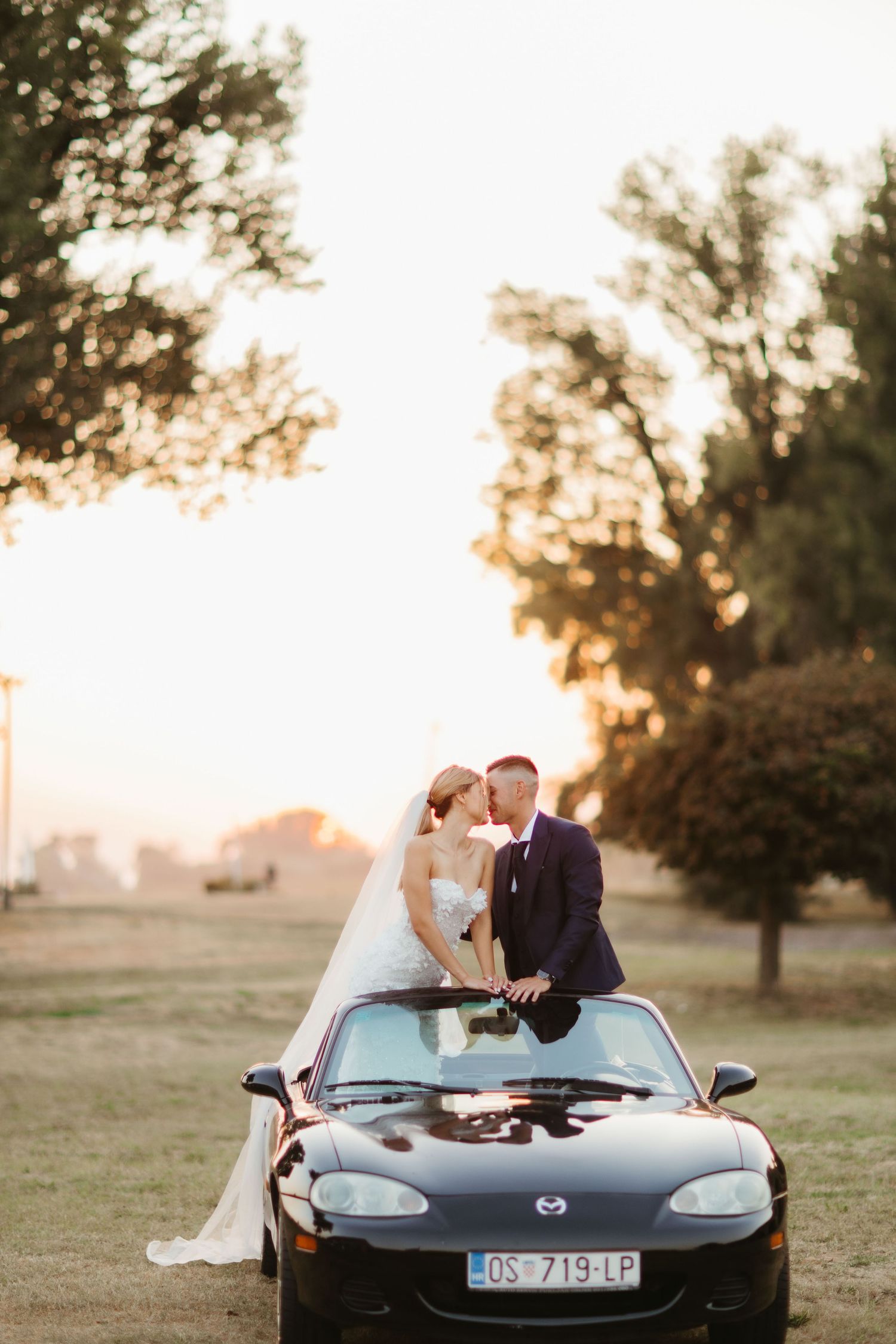 Romantic sunset wedding photo with couple posing on black convertible sports car.