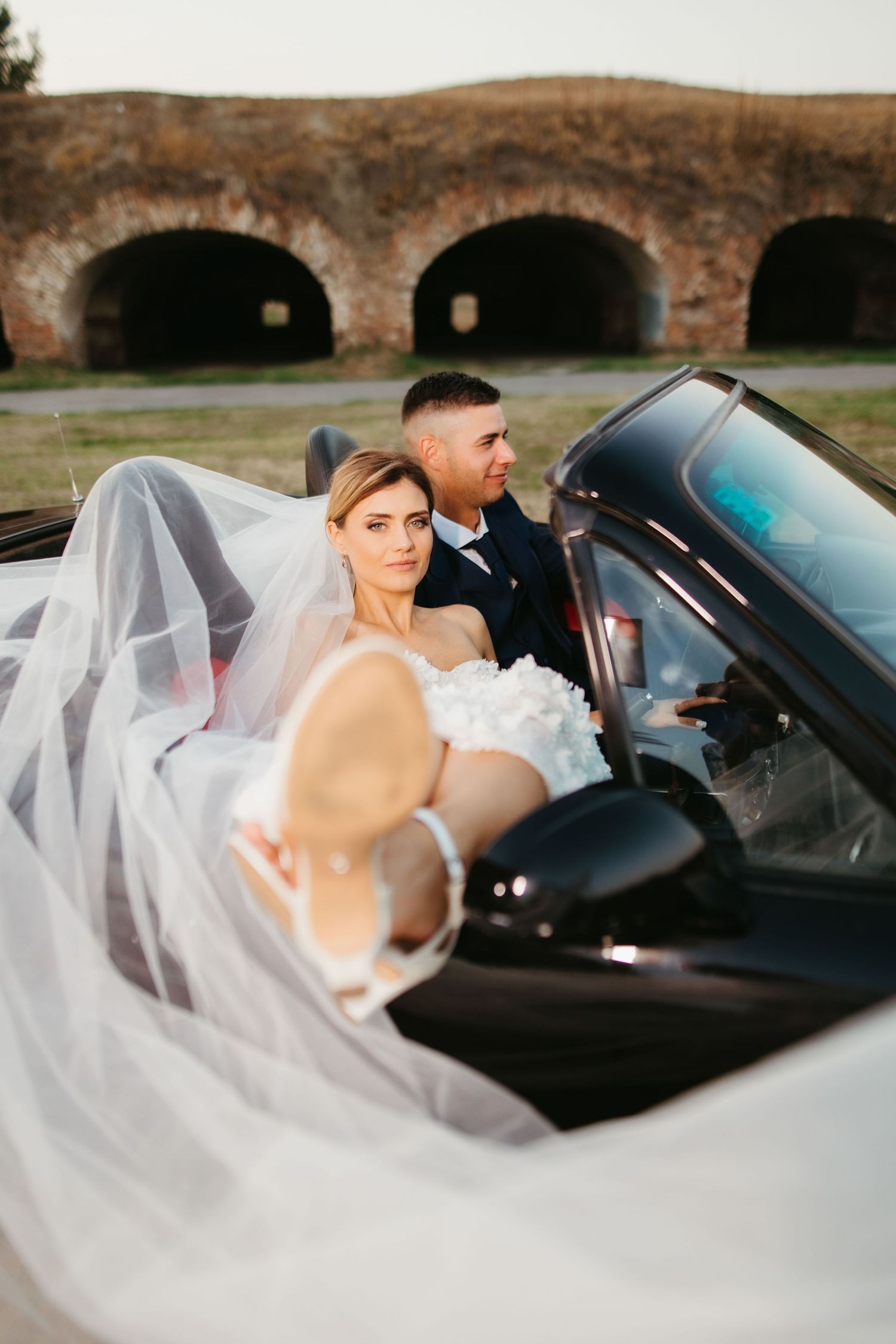 Wedding couple in convertible with stone archways in background.