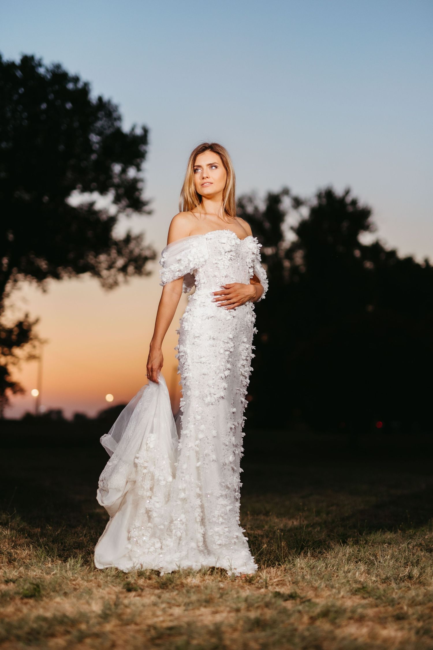 A full-length portrait of a bridal gown with intricate floral details against a sunset backdrop.