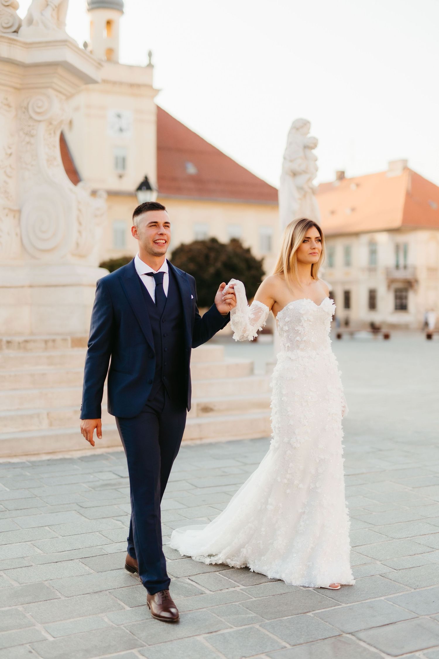 Bride in lace gown and groom in navy suit stroll through historic plaza.
