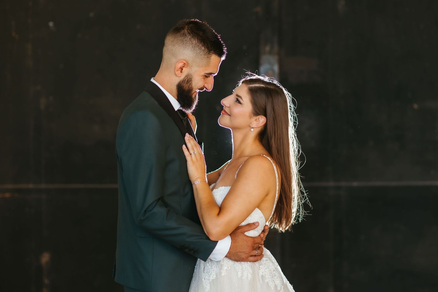 Newlywed couple embraces intimately in an elegant wedding photo with dramatic dark background lighting.