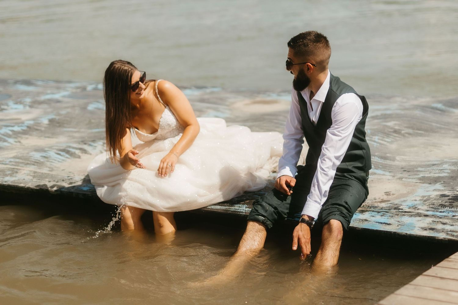 A couple sits at the ocean's edge in formal attire, letting the waves splash around them during a romantic photoshoot.
