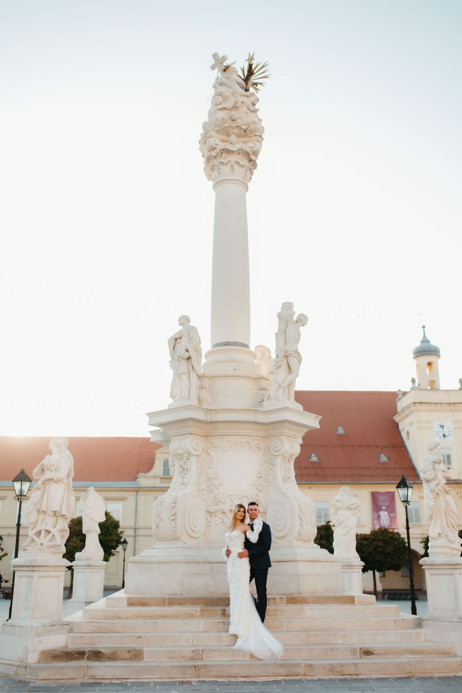 Wedding couple poses at base of ornate white stone monument during golden hour.