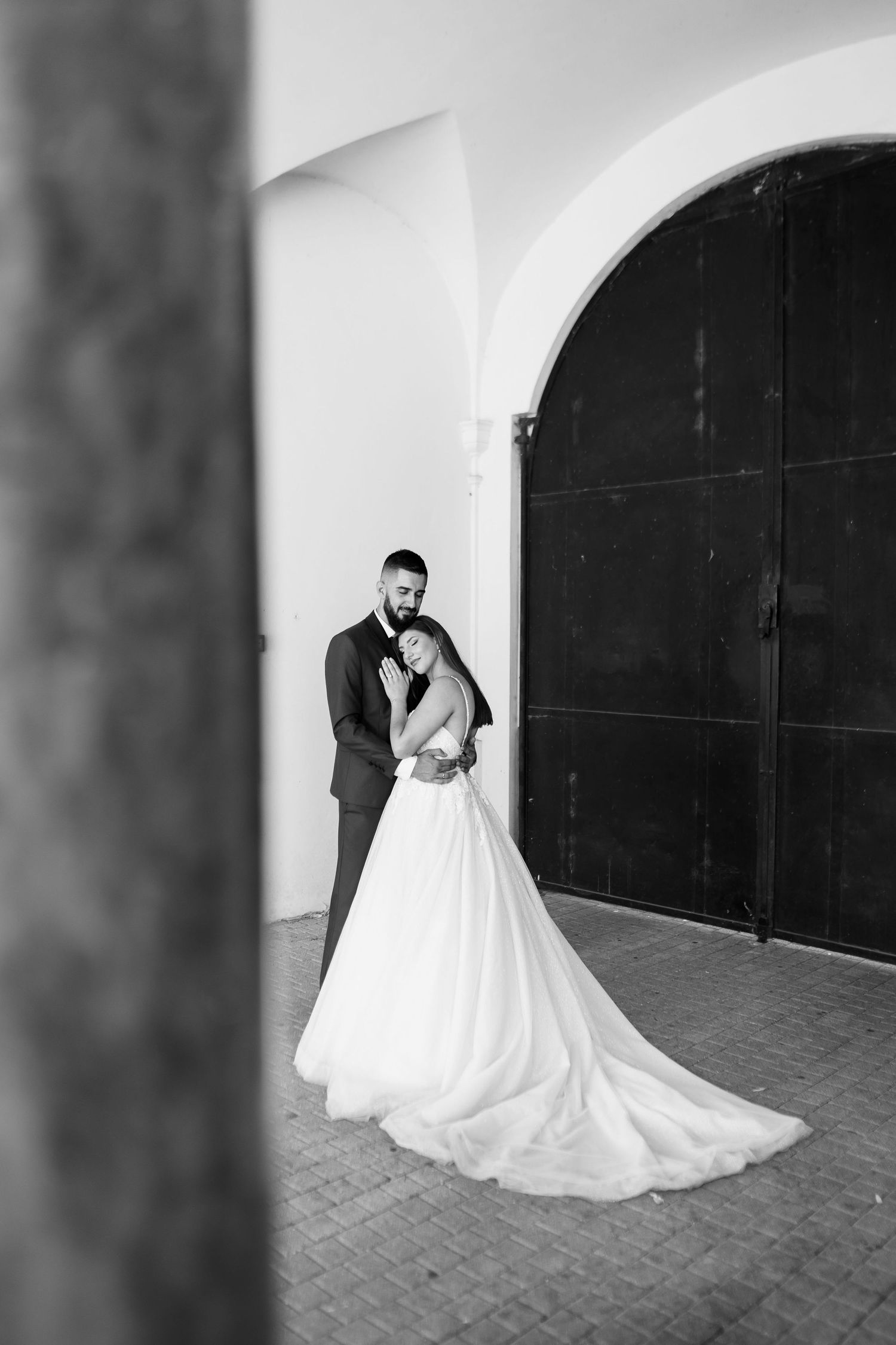 A romantic black and white wedding portrait showing a couple embracing in front of a large arched doorway.