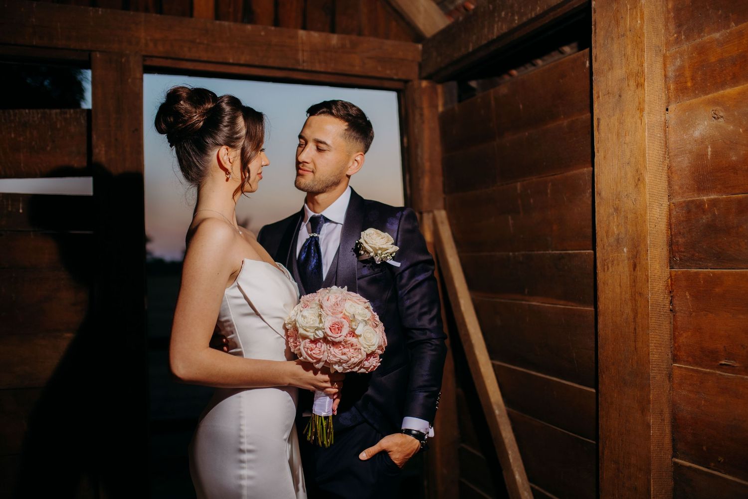 Newlyweds share an intimate moment in a rustic wooden barn setting illuminated by warm lighting.