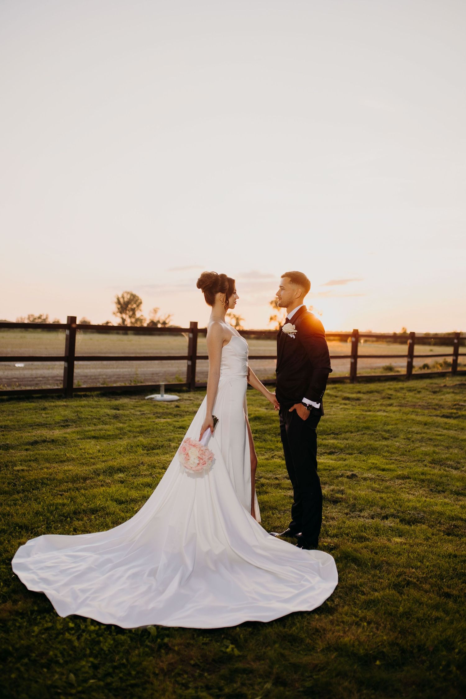 Elegant silhouette captures a couple's romantic wedding moment against a sunset in a rustic farm setting with wooden fencing.