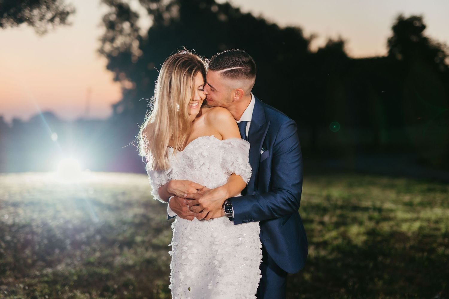 Couple shares romantic kiss at dusk in elegant wedding attire against natural outdoor setting.